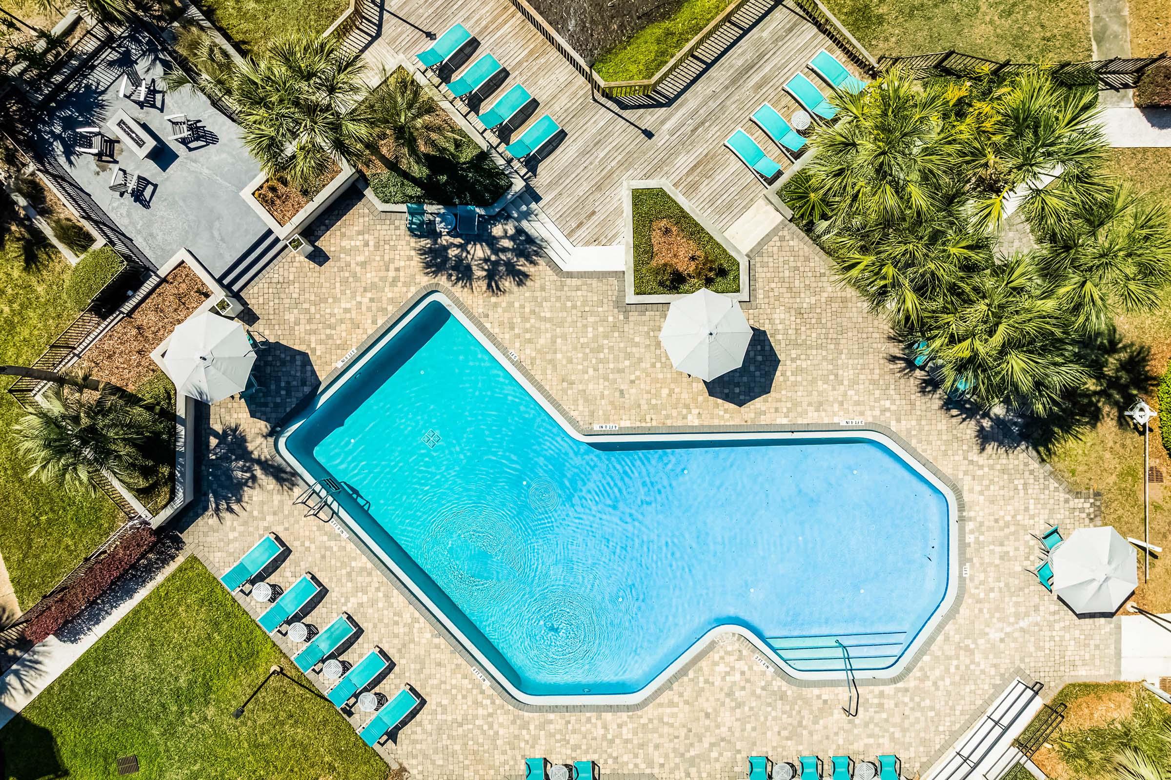 Aerial view of a swimming pool shaped like a guitar, surrounded by lounge chairs and umbrellas. The pool area is landscaped with greenery, featuring wooden decking and a relaxing atmosphere perfect for outdoor leisure.