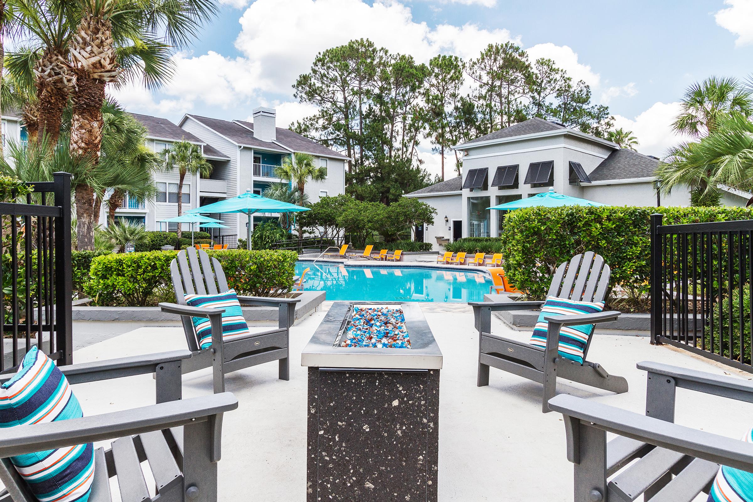 A vibrant outdoor pool area surrounded by lush greenery. Comfortable lounge chairs in bright colors are positioned around the pool, with tables and umbrellas providing shade. In the foreground, wooden adirondack chairs face a fire pit, creating a cozy atmosphere for relaxation.