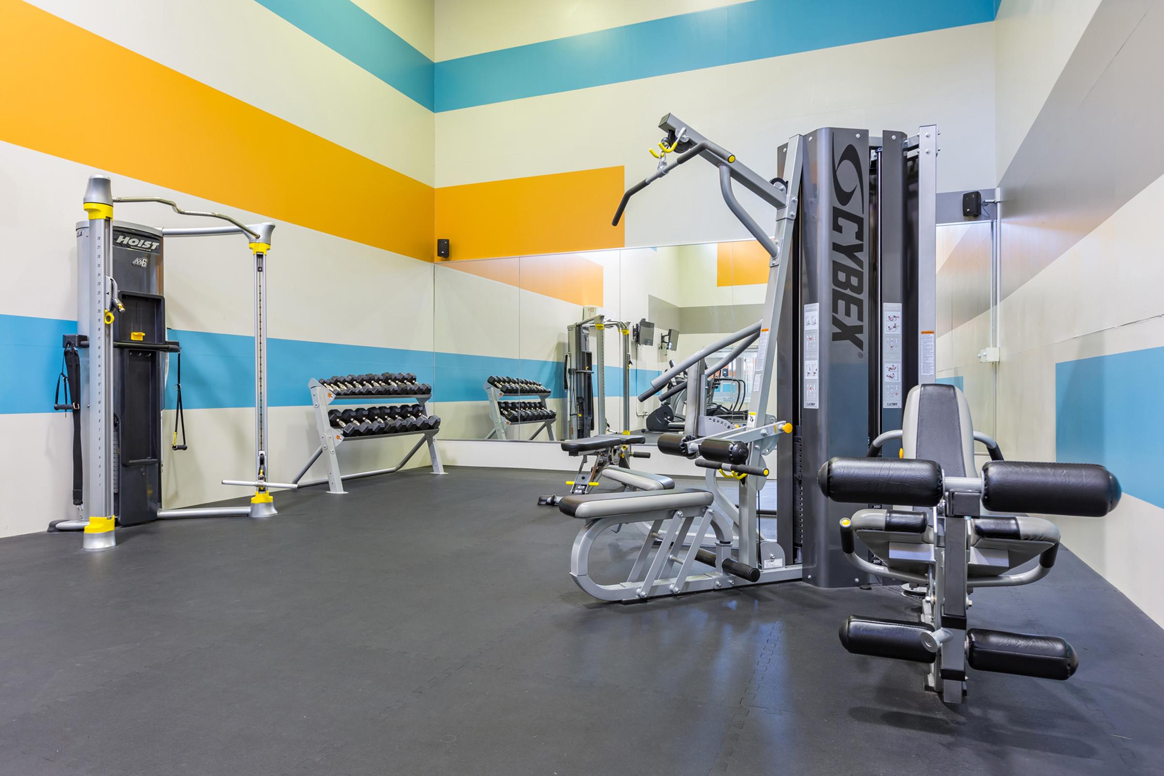 A modern gym interior featuring a variety of fitness equipment. The space includes a multi-station machine, free weights on racks, and a mirrored wall. The walls are painted with colorful horizontal stripes in blue, orange, and yellow, creating a vibrant atmosphere. The flooring is dark and smooth, suitable for exercise.