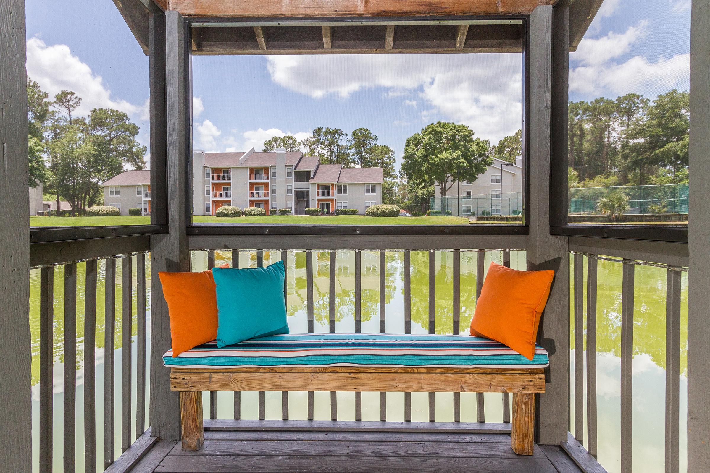 A cozy wooden seating area with teal and orange cushions overlooks a serene pond surrounded by greenery and residential buildings. The peaceful setting is complemented by a clear blue sky with fluffy clouds reflecting in the water.