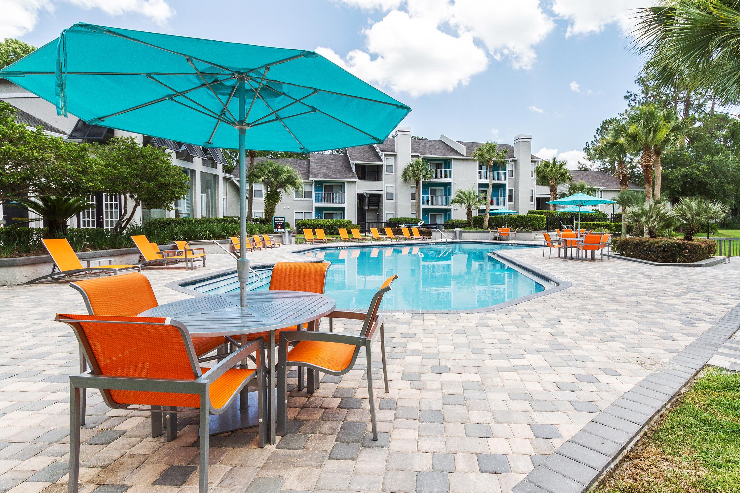 A vibrant outdoor pool area featuring lounge chairs and tables with orange cushions, surrounded by lush greenery. Large teal umbrellas provide shade over the dining areas, and the serene pool reflects a clear blue sky with fluffy clouds. Elegant apartment buildings are visible in the background.