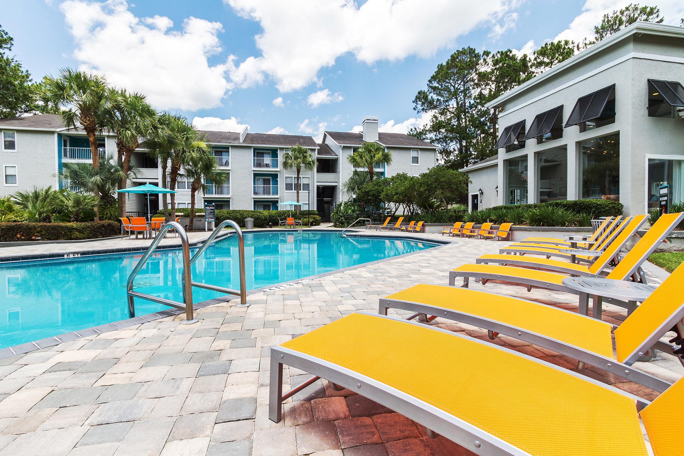 A sunny pool area featuring a blue swimming pool surrounded by vibrant orange lounge chairs. Lush greenery and palm trees provide a tropical atmosphere, while modern apartment buildings and bright clouds complete the scenic view. Perfect for relaxation and leisure activities.