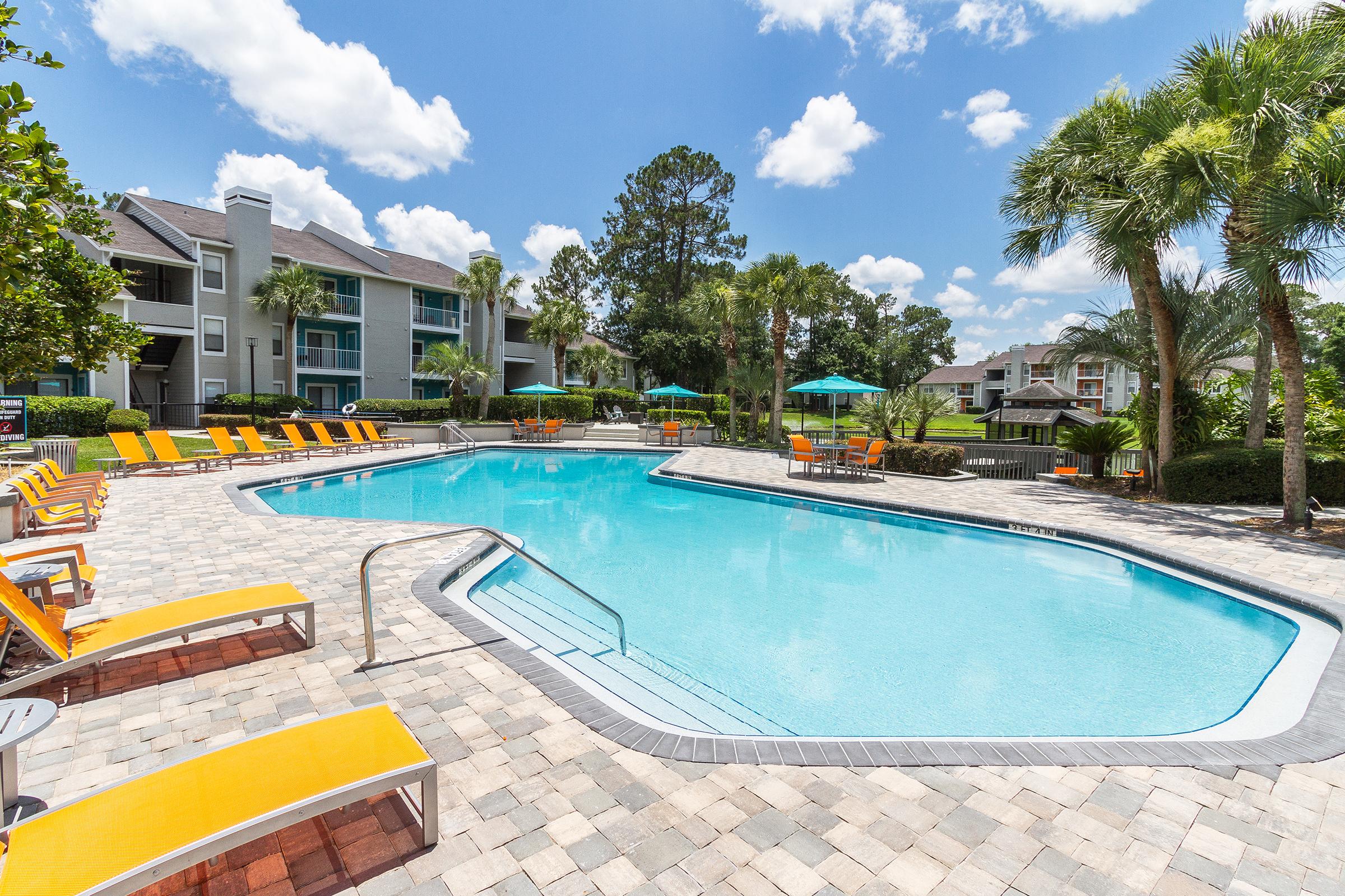 A clear blue swimming pool surrounded by orange lounge chairs and palm trees. Apartment buildings are visible in the background under a bright blue sky with white clouds, creating a relaxing outdoor setting.