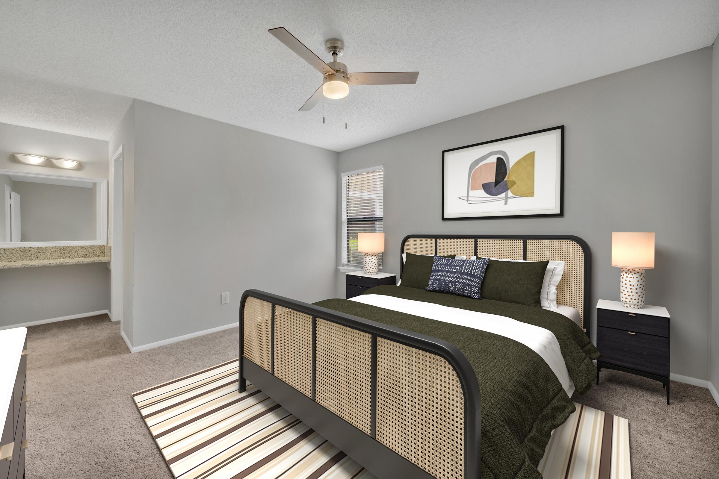 A modern bedroom featuring a sleek bed with a dark green and white bedding set. There are two nightstands with lamps on either side, and a decorative wall art piece above the bed. A ceiling fan provides lighting, and a striped area rug adds texture to the carpeted floor. A mirror and countertop are visible in the adjoining area.