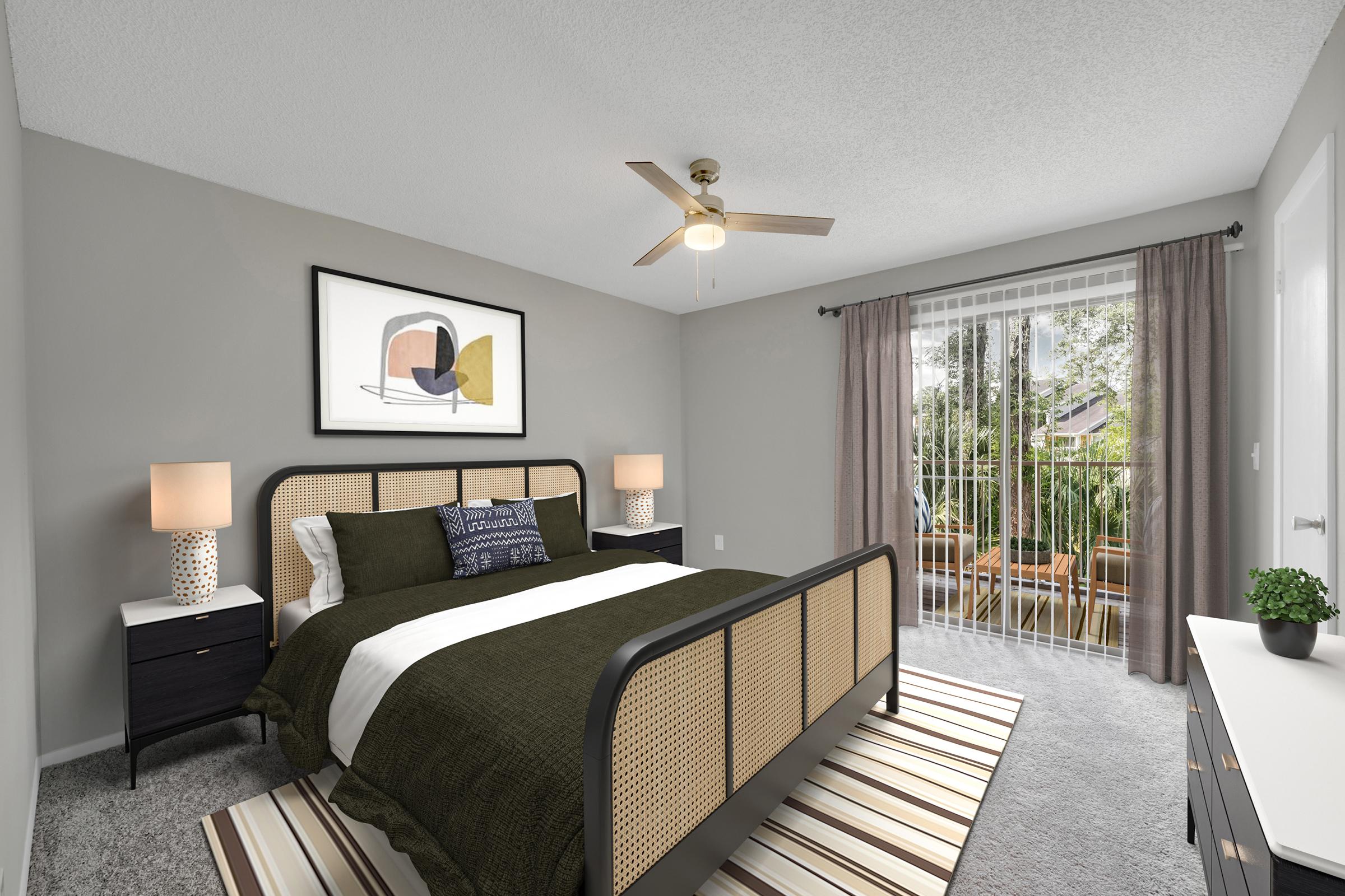 A modern bedroom featuring a stylish bed with a dark green and white bedding set. There are two bedside lamps on matching nightstands, a wall art piece above the bed, and a window with sheer curtains leading to a patio. A ceiling fan adds to the cozy atmosphere with gray walls and carpet.