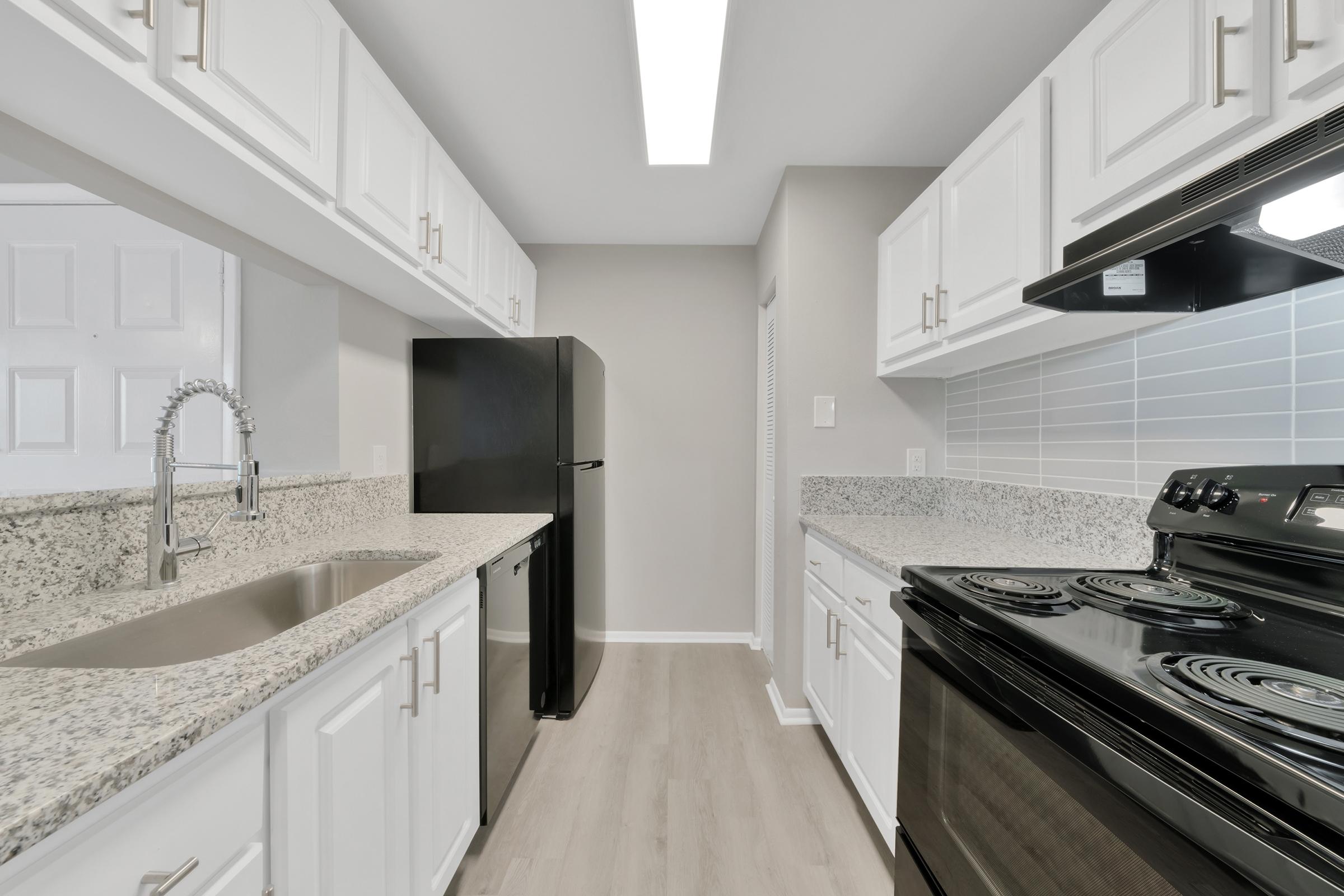 A modern kitchen featuring white cabinetry, granite countertops, a black refrigerator, and a black stove. The space is well-lit with a ceiling light and has light-colored flooring.