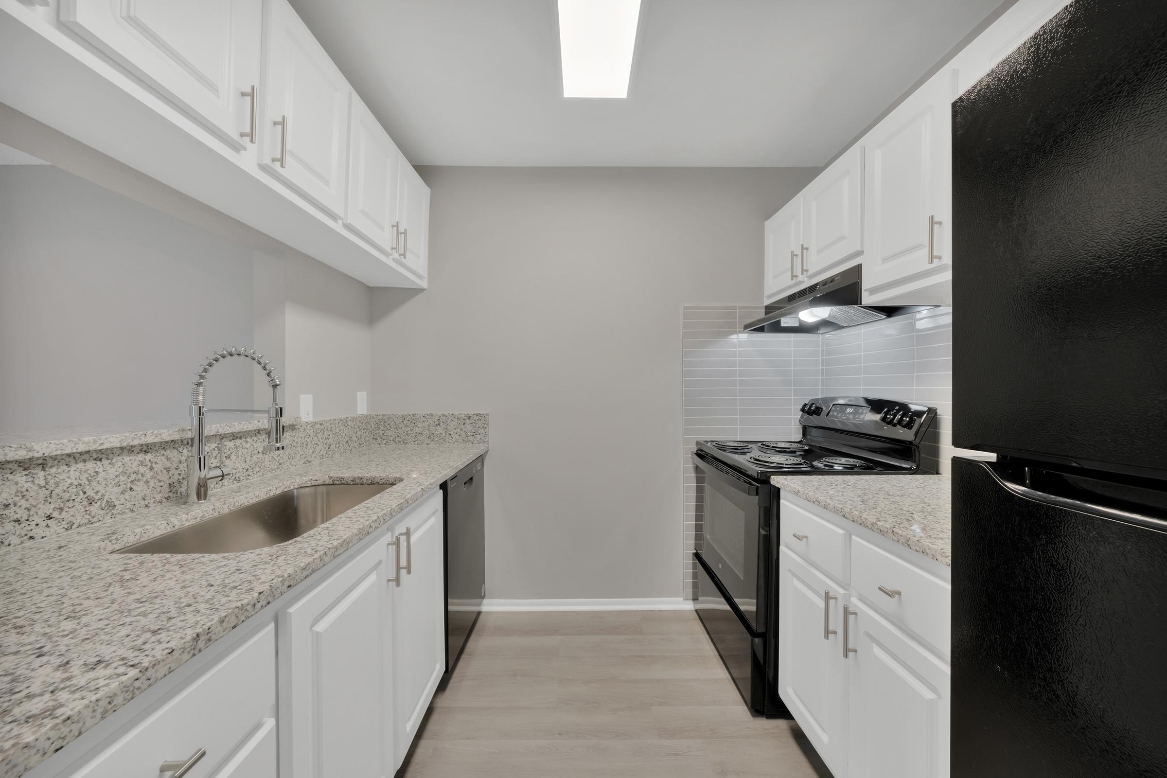 Modern kitchen featuring granite countertops, white cabinetry, a stainless steel sink with a pull-down faucet, a black refrigerator, and a black gas stove. Bright lighting from the ceiling fixture illuminates the space, with a light gray wall providing a neutral backdrop.