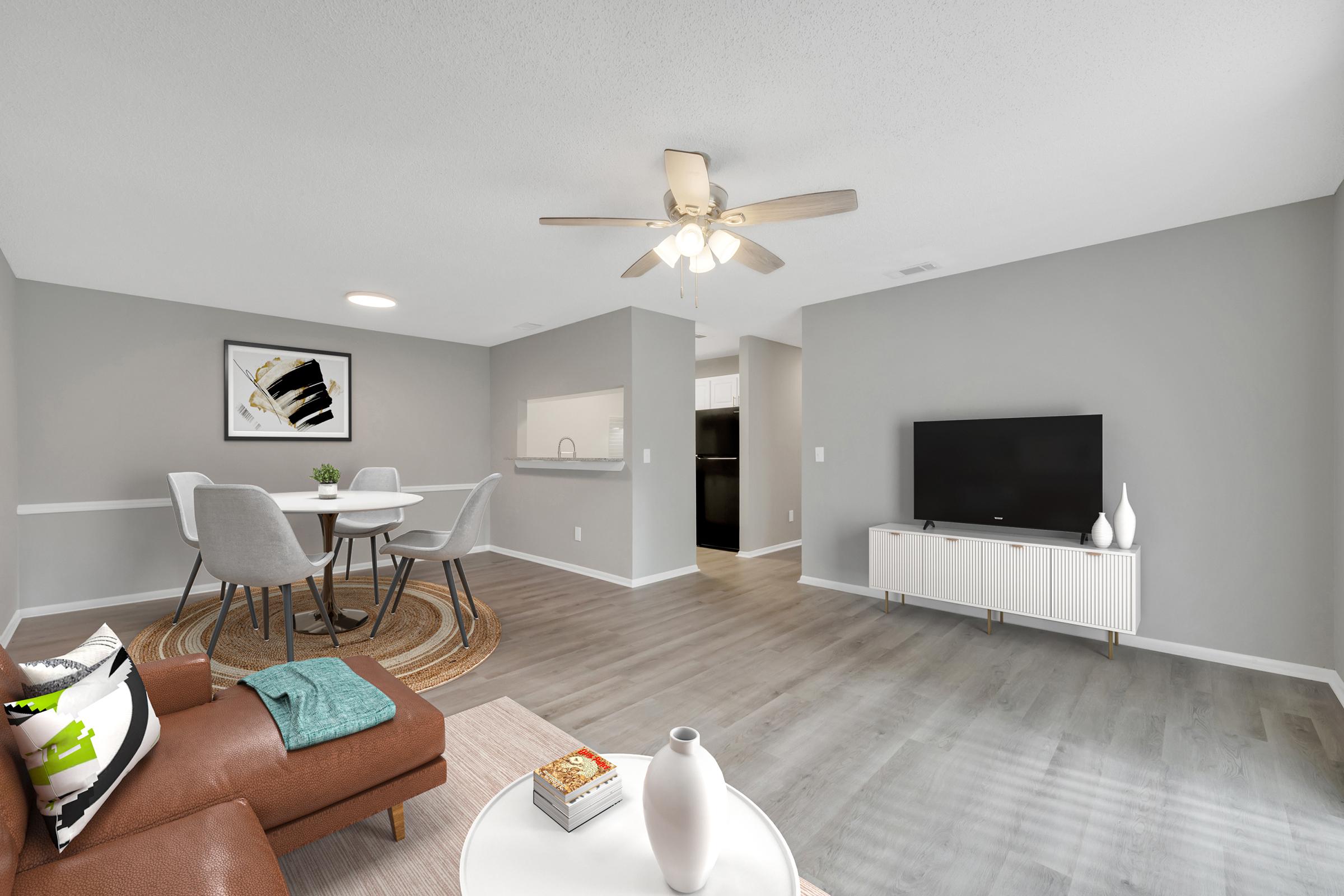 A modern living room featuring a comfortable brown sofa, a round white coffee table, and a neutral color scheme. A dining area with a round table and gray chairs is visible. A ceiling fan hangs above, and a large flat-screen TV is mounted on the wall. The kitchen area is partially visible in the background.