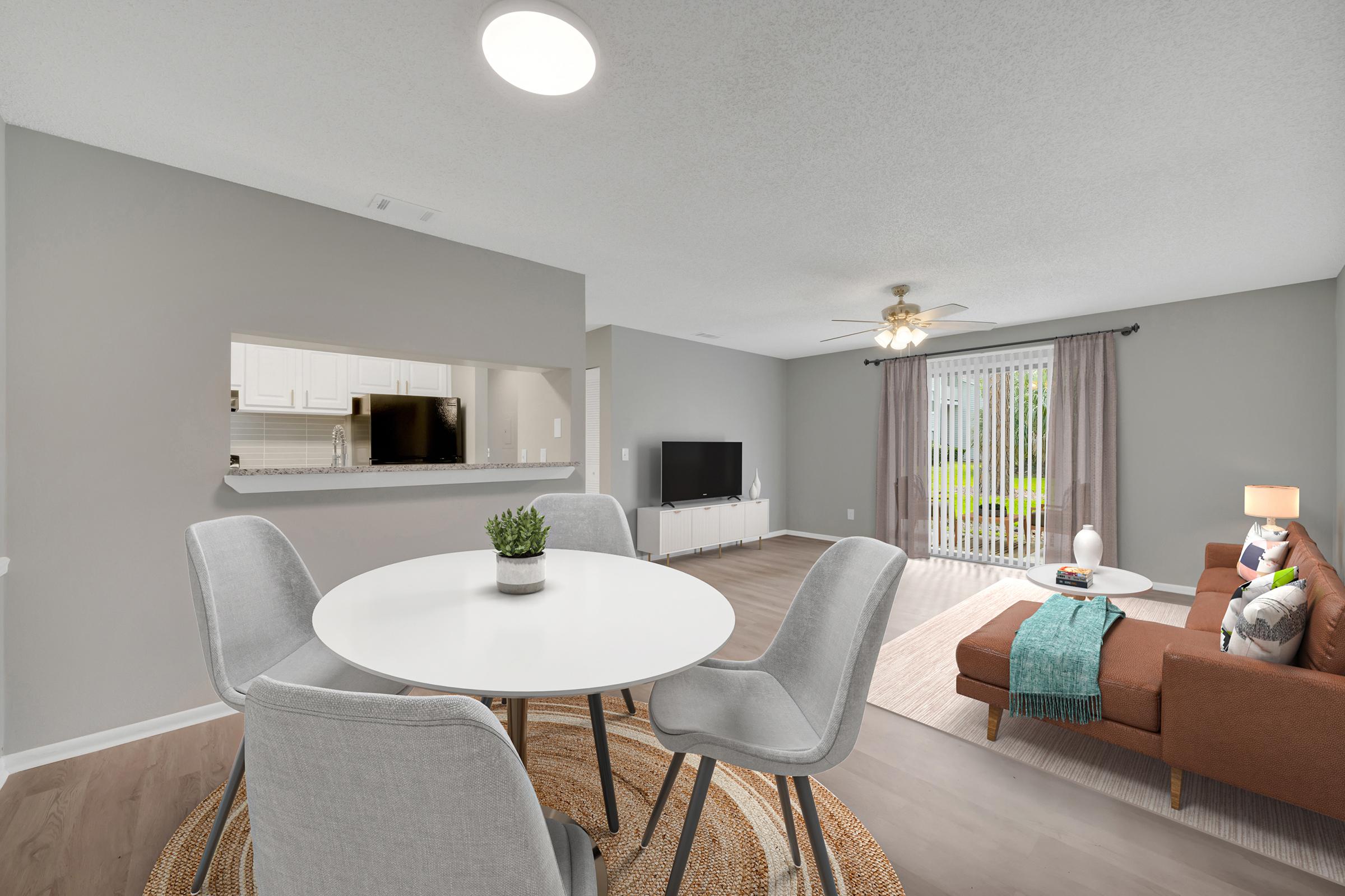 A modern living space featuring a round white dining table with four gray chairs, a brown couch with decorative pillows, and a large window with curtains. There is a TV on a white media console, and a cozy rug on the floor. The room has light gray walls and bright lighting, creating an inviting atmosphere.