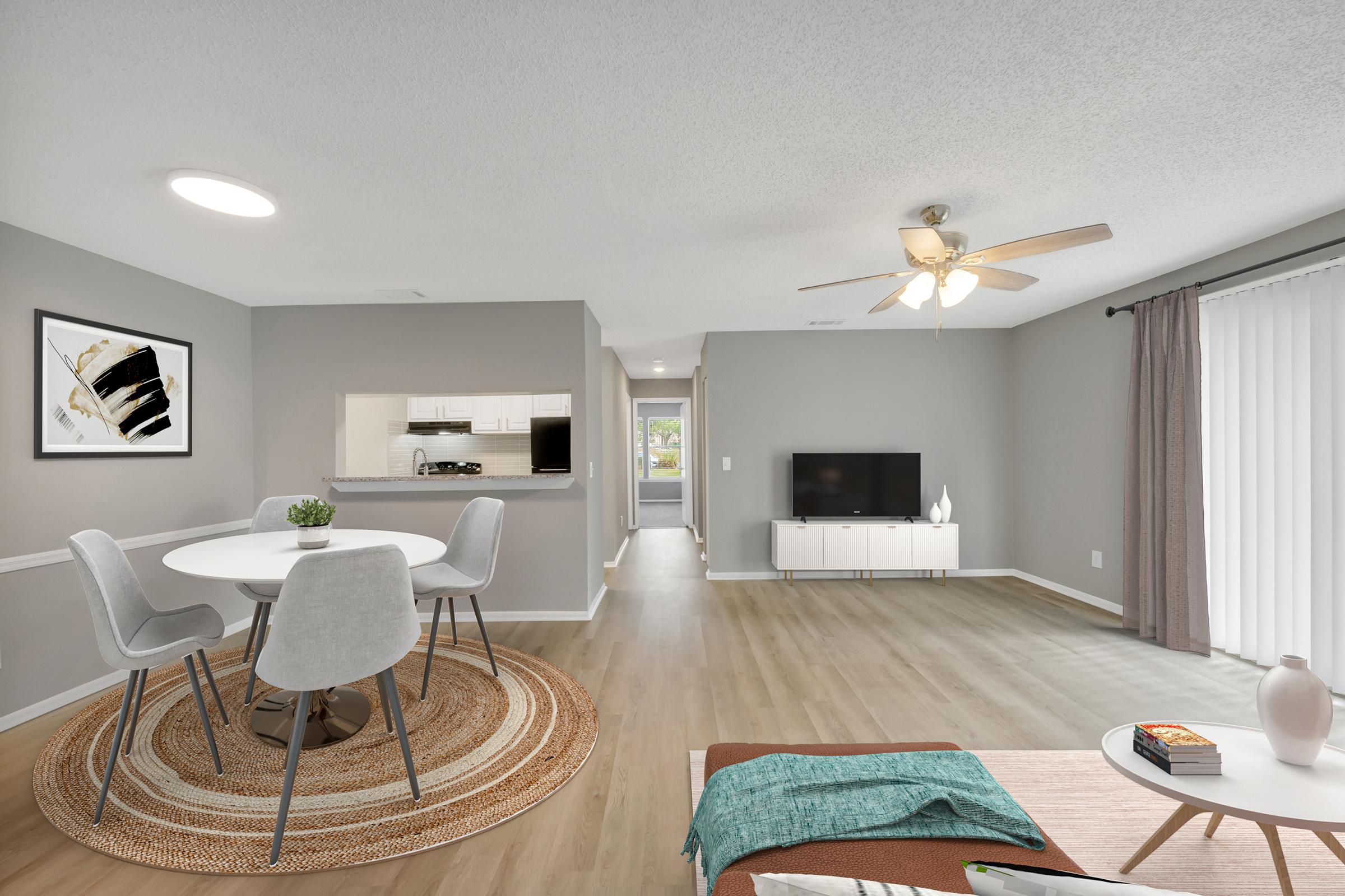 Bright and airy living space featuring a round dining table with gray chairs, a modern TV stand, and a cozy sitting area with a coffee table. The walls are painted in light gray, with a textured rug on the floor. A ceiling fan provides a comfortable atmosphere, and large windows let in natural light.