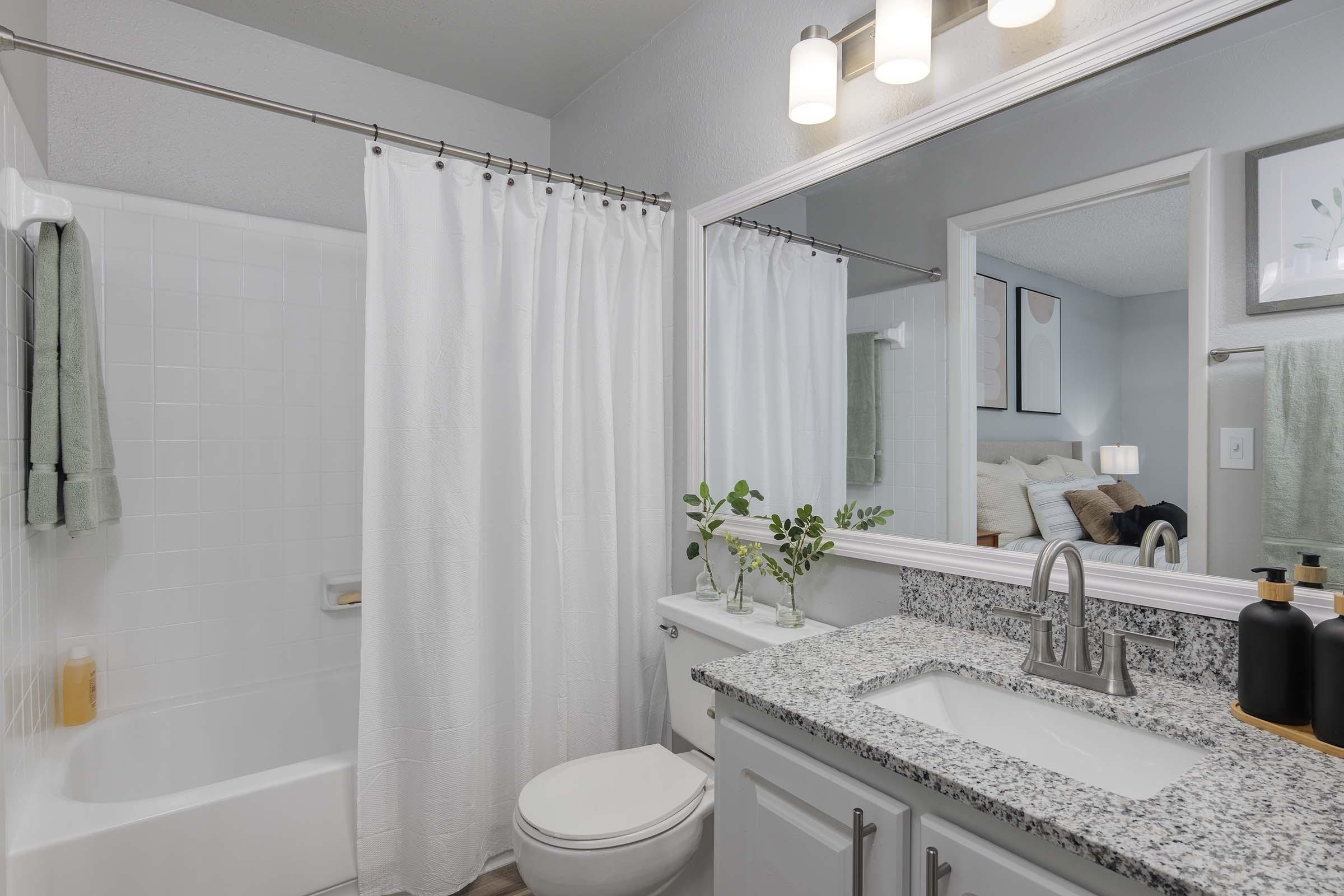 A modern bathroom featuring a bathtub with a shower curtain, a granite countertop with a sink, a mirror above the sink, and decorative plants. The walls are light-colored, and a glimpse of a bedroom is visible through the doorway in the background. Soft lighting fixtures illuminate the space.