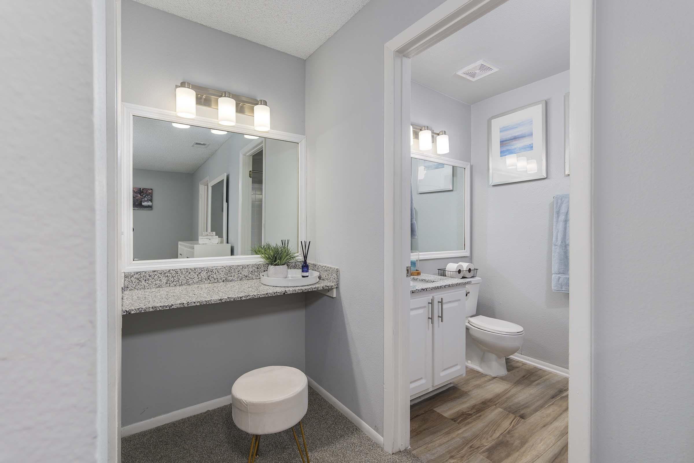 A modern bathroom featuring a double vanity with a light mirror above, a white toilet, and a small cushioned stool. The walls are painted light gray, and there is a decorative plant on the vanity. The floor has wood-like tiles, and another room is visible in the background.