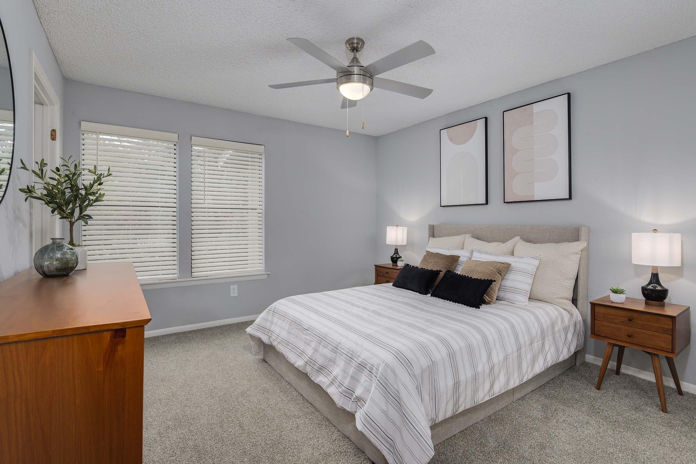 A modern bedroom featuring a beige bed with white and gray striped bedding. There are two bedside tables with lamps on either side and a wooden dresser against the wall. The room has light gray walls, two windows with blinds, and abstract art on the wall above the bed. A ceiling fan is also visible.