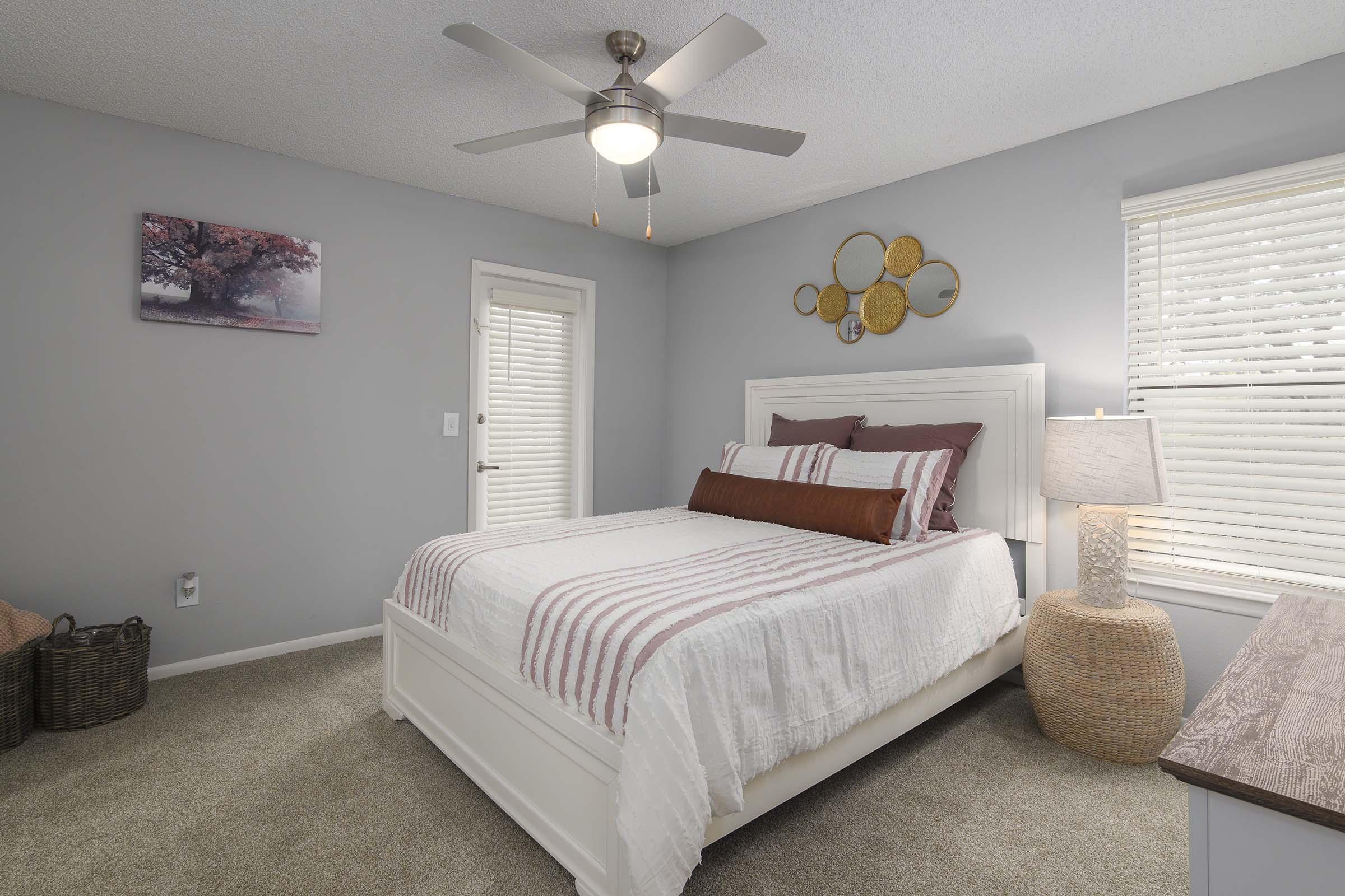 A cozy bedroom featuring a white bed with striped bedding, a decorative throw pillow, and a bedside lamp. The walls are painted light gray, adorned with a framed picture of a tree, and a round mirror with a sunburst design. A door leads to another room, and the carpet is plush and neutral-toned.