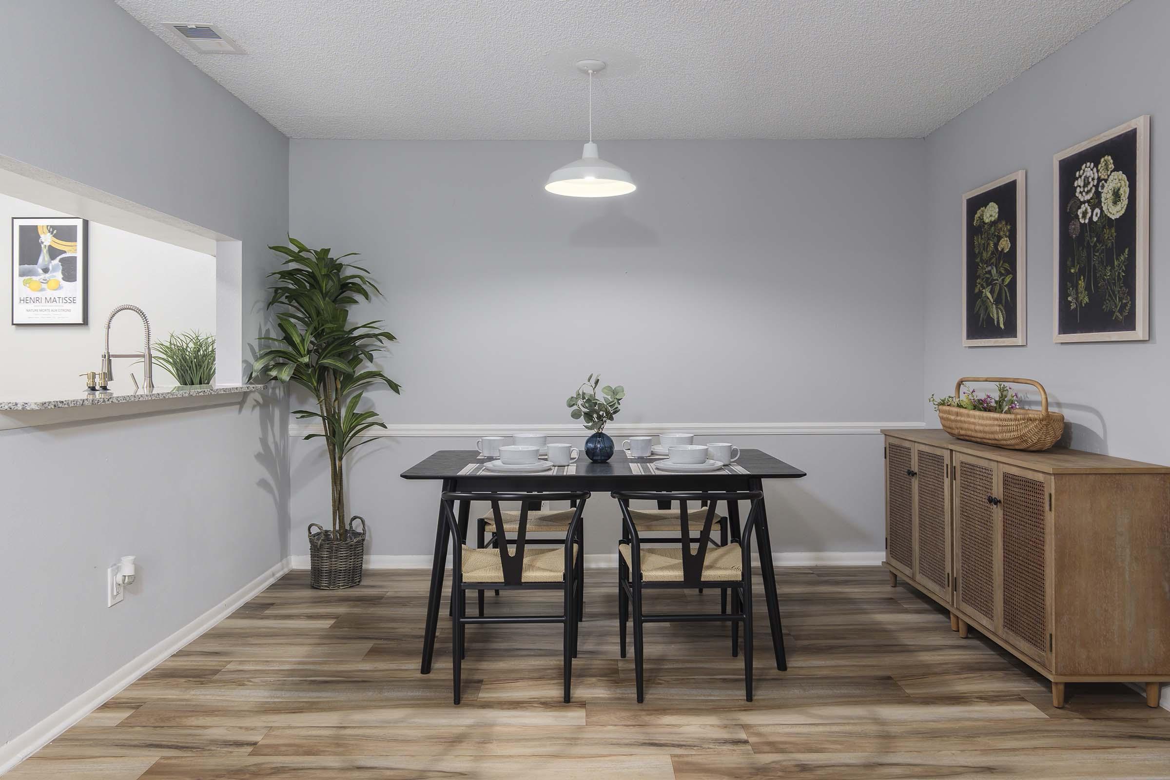 A modern dining area with a black table and chairs set for a meal, featuring white dishes and a blue vase. A light pendant hangs above the table. To the left, a kitchen bar with plants can be seen, while the right displays a wooden sideboard and two framed floral artworks on the pale gray wall.