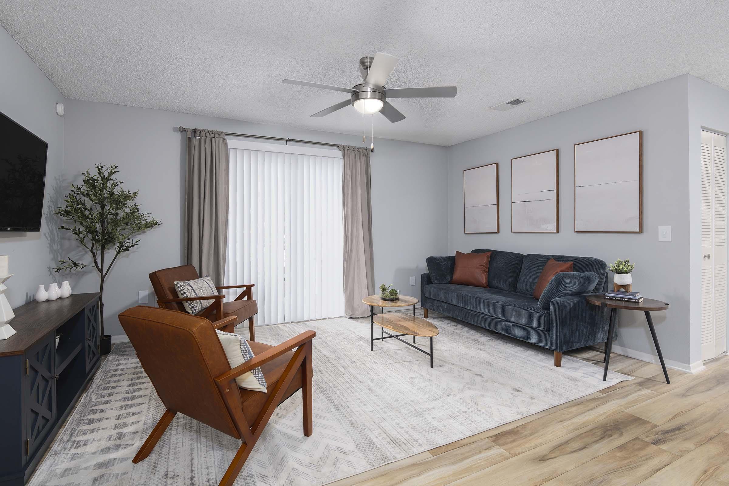 A modern living room featuring a navy blue sofa, two brown armchairs, and a wooden coffee table on a light-colored rug. Natural light filters through sheer curtains, and there are three framed pictures on the wall. A TV is mounted on a dark blue media console, complemented by a decorative plant in the corner.