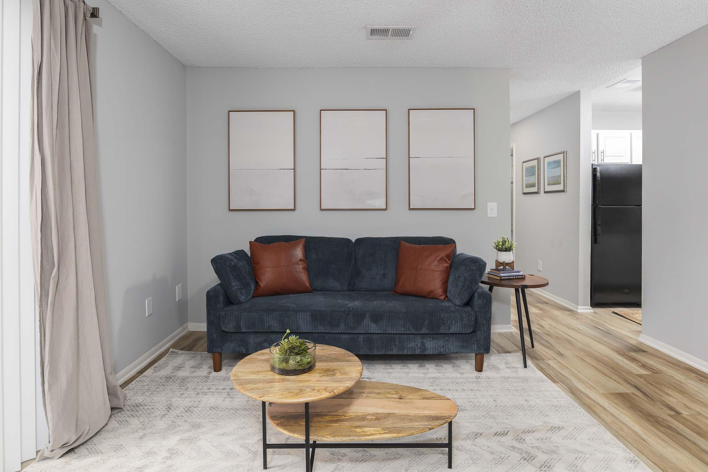 A cozy living room featuring a blue sofa with two orange cushions. There are three abstract art pieces on the wall and a round wooden coffee table with a plant. A rug covers the floor, and the space is lit by natural light, with a view of a kitchen in the background.