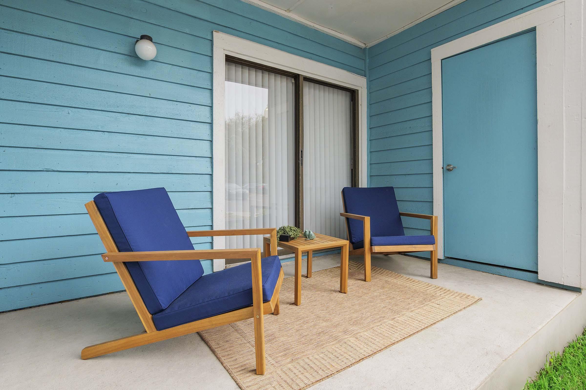 A cozy porch area featuring two blue chairs and a small wooden table on a light-colored rug. The walls are painted a bright blue, and there is a glass door with vertical blinds, as well as a blue door on the side.