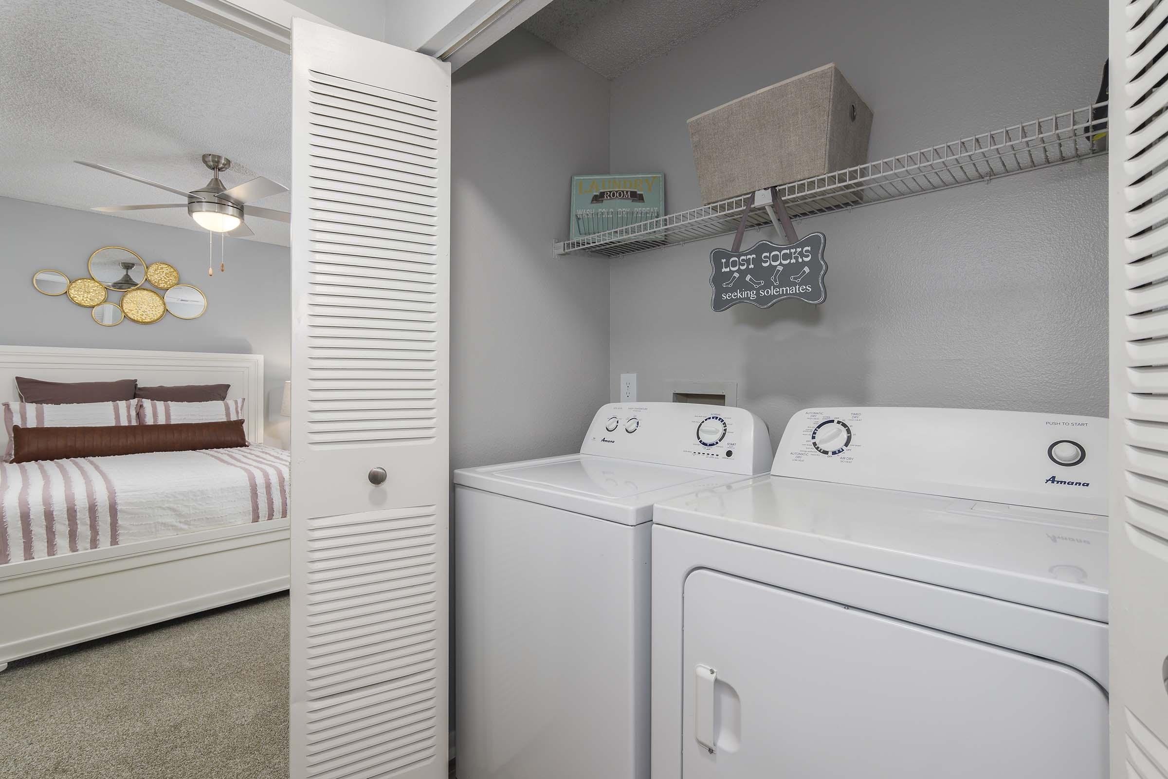 A laundry room featuring a washer and dryer, with a wall-mounted shelf above. To the side, there is a door leading to a bedroom with a bed adorned in white and red striped bedding. The room is well-lit and includes decorative items, such as a sign reading "Lost Socks."
