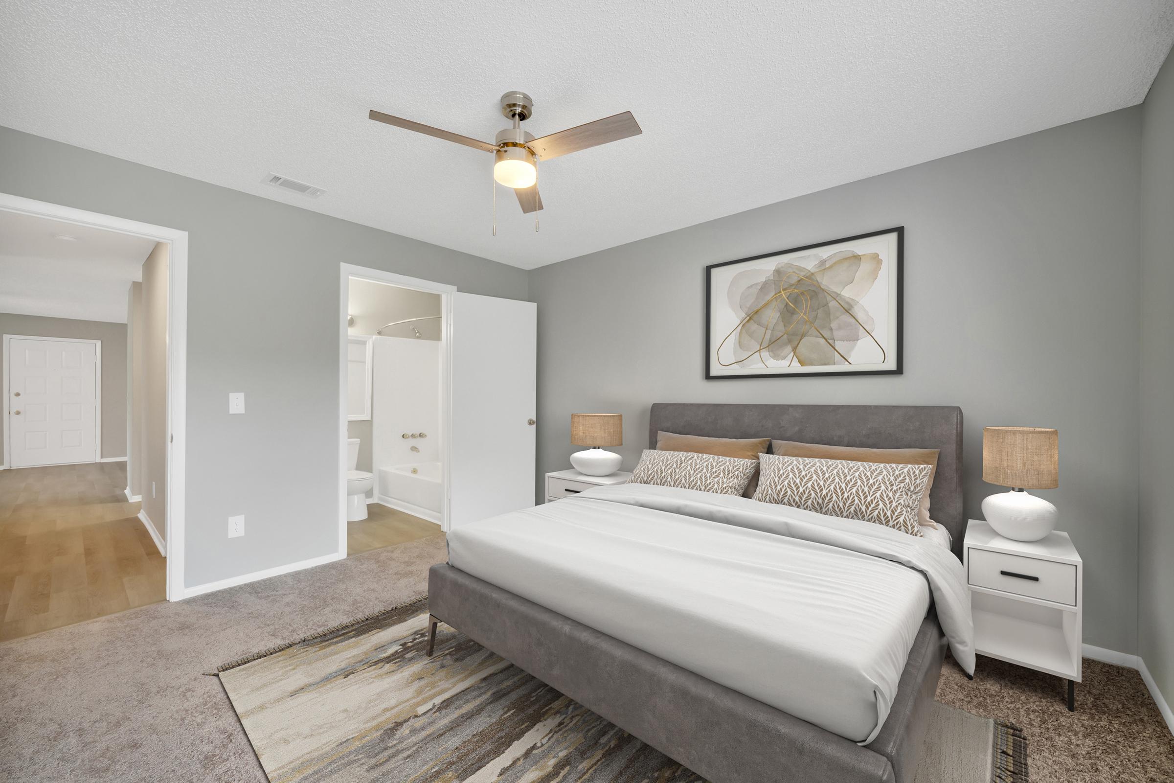 A modern bedroom featuring a gray color scheme, a comfortable bed with decorative pillows, two stylish nightstands with lamps, and a ceiling fan. An open door reveals a bathroom area. The room has a minimalist aesthetic with a rug and artwork on the wall, creating a serene atmosphere.
