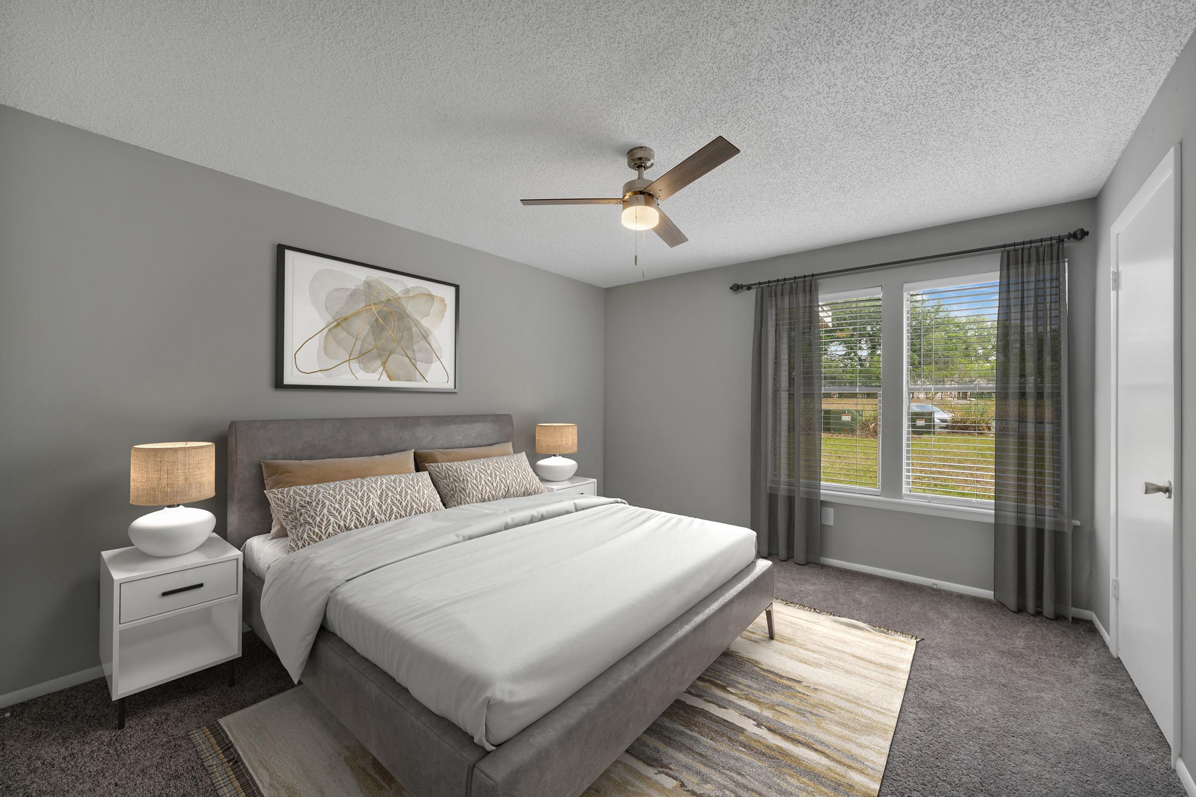 A modern bedroom featuring a gray accent wall, a queen-sized bed with white bedding, and decorative pillows. Two bedside tables with lamps are on either side of the bed. A ceiling fan hangs above, and a large window with blinds lets in natural light, offering a view of the outdoors. A soft area rug completes the decor.