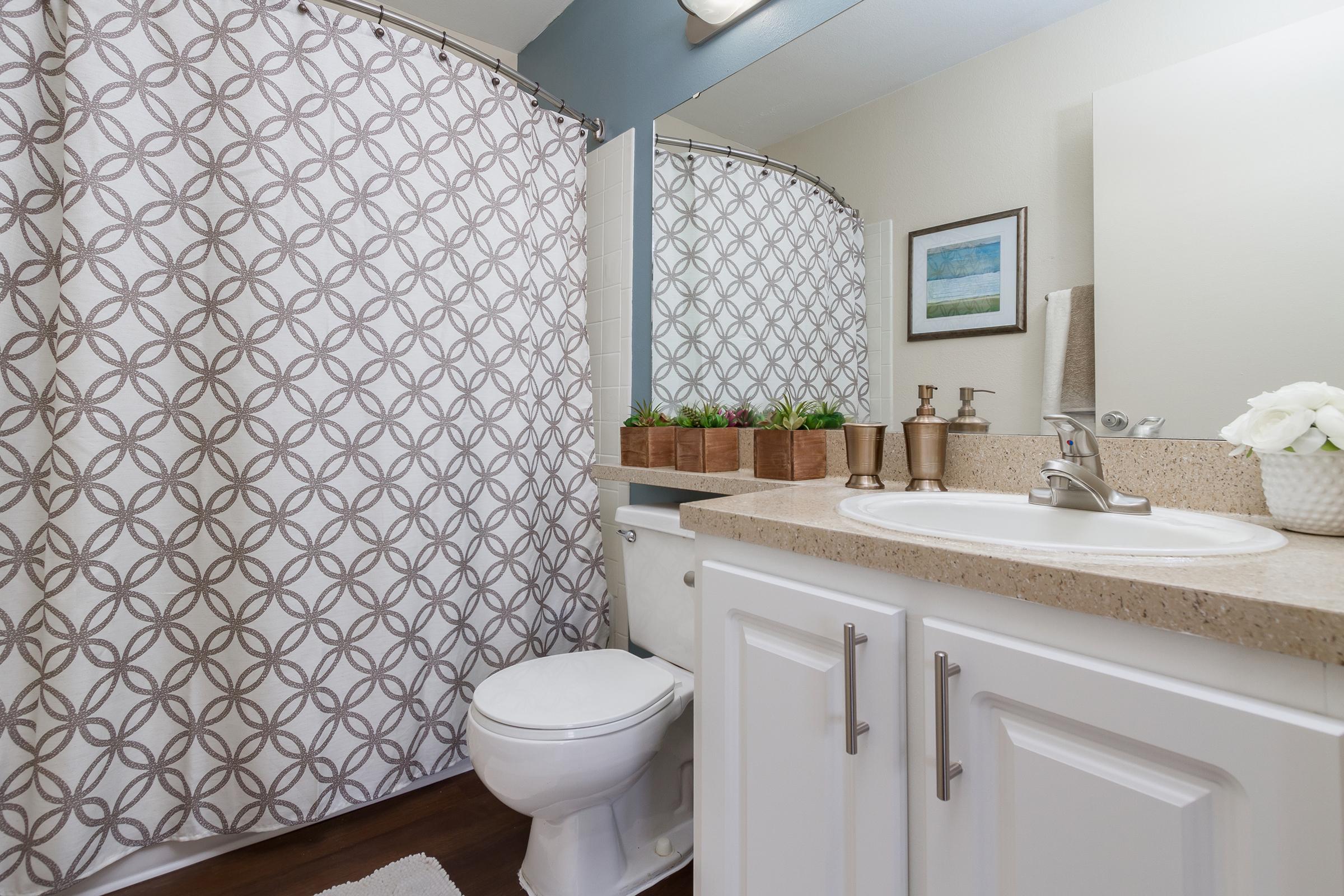 A bright and modern bathroom featuring a white toilet, a sink with a sleek faucet, and a countertop. The shower is enclosed with a patterned curtain. Above the sink, there is a mirror and a small framed artwork. Green plants are arranged in two decorative containers on the countertop, adding a touch of nature.