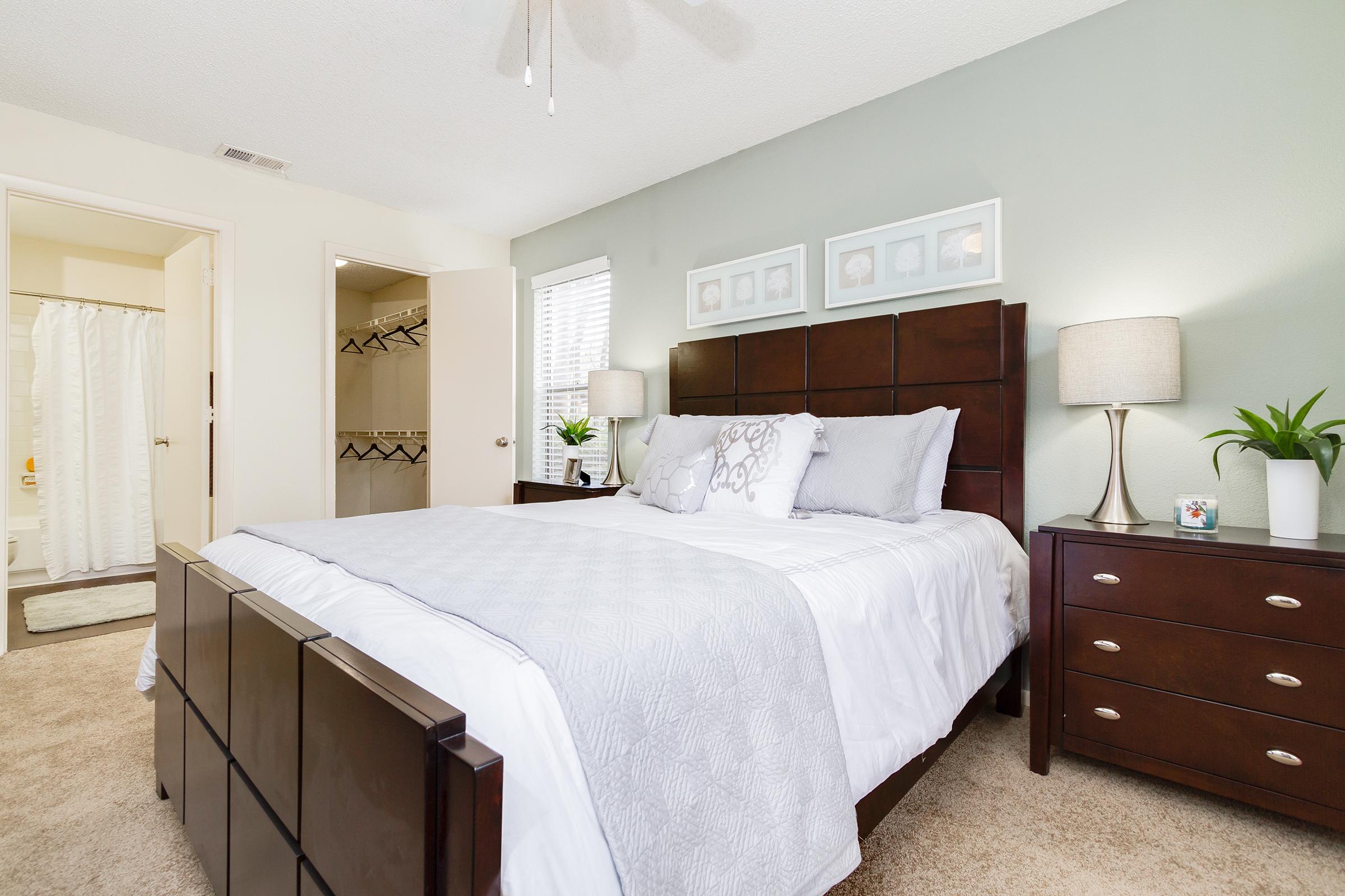 A cozy bedroom featuring a queen-sized bed with gray and white bedding, two bedside lamps on matching dressers, and framed artwork above the headboard. A doorway leads to a bathroom with a shower curtain, and a closet with hanging space is visible. The walls are painted a soft green, enhancing the tranquil atmosphere.