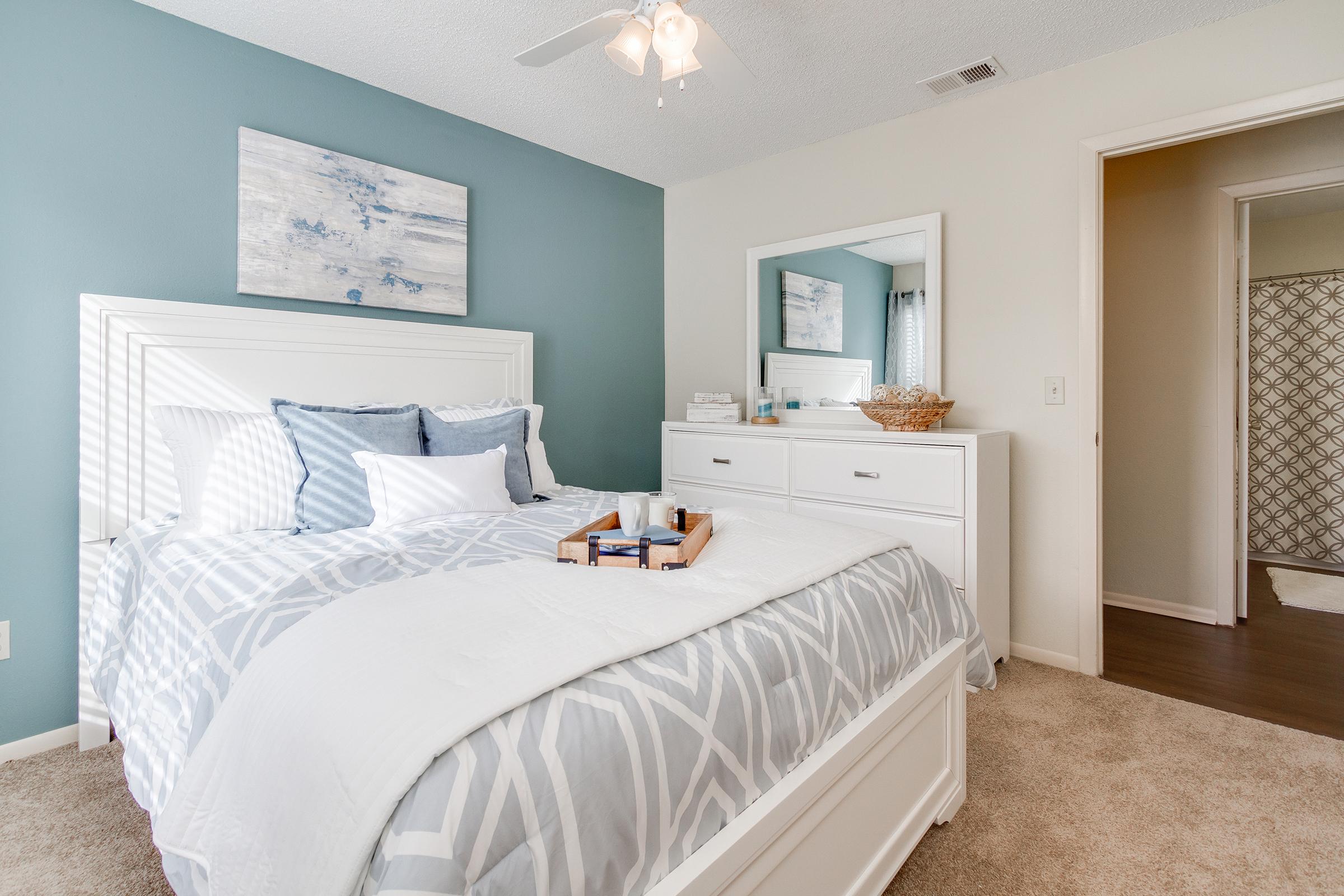 A cozy bedroom featuring a white bed with patterned bedding, accented by blue pillows. There's a nightstand with a tray and books, a large mirror, and a dresser. Soft lighting from a ceiling fan creates a welcoming atmosphere. A bathroom door is visible in the background.
