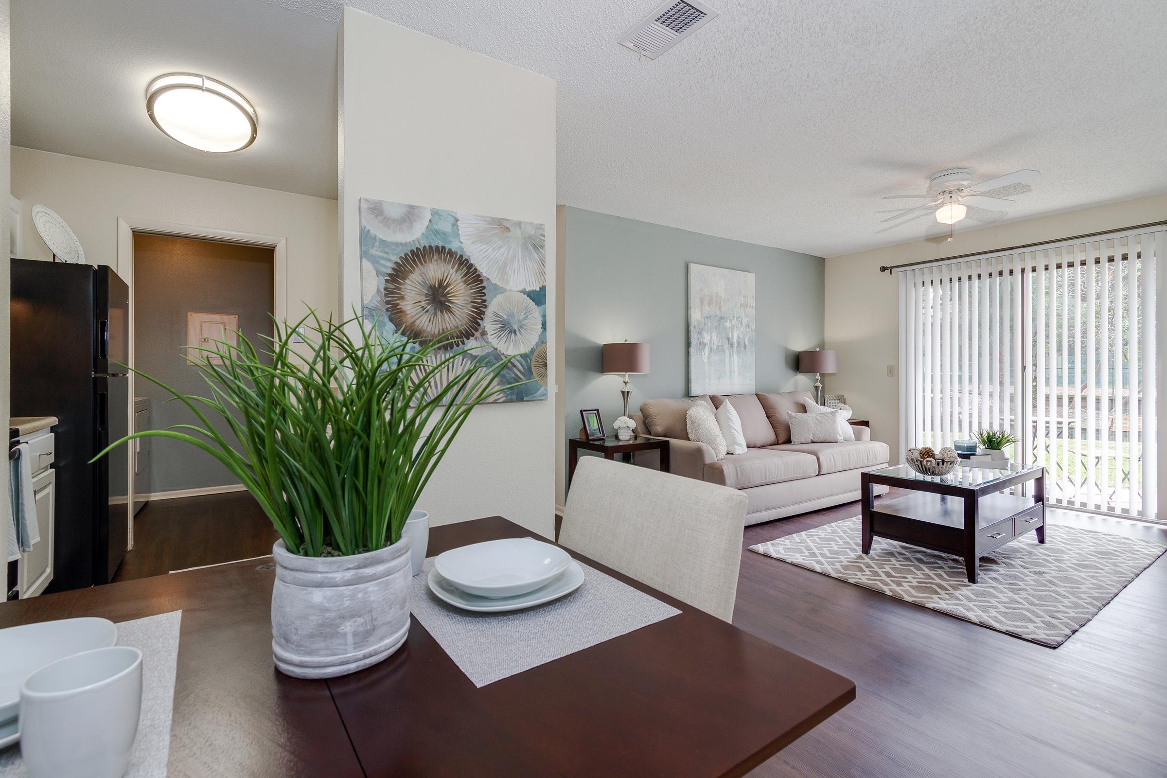 A cozy living space featuring a light-colored sofa, a wooden coffee table, and a ceiling fan. The room has large windows with white blinds, allowing natural light to enter. In the foreground, a dining table is set with plates and a plant centerpiece. The decor is modern and inviting, creating a warm atmosphere.