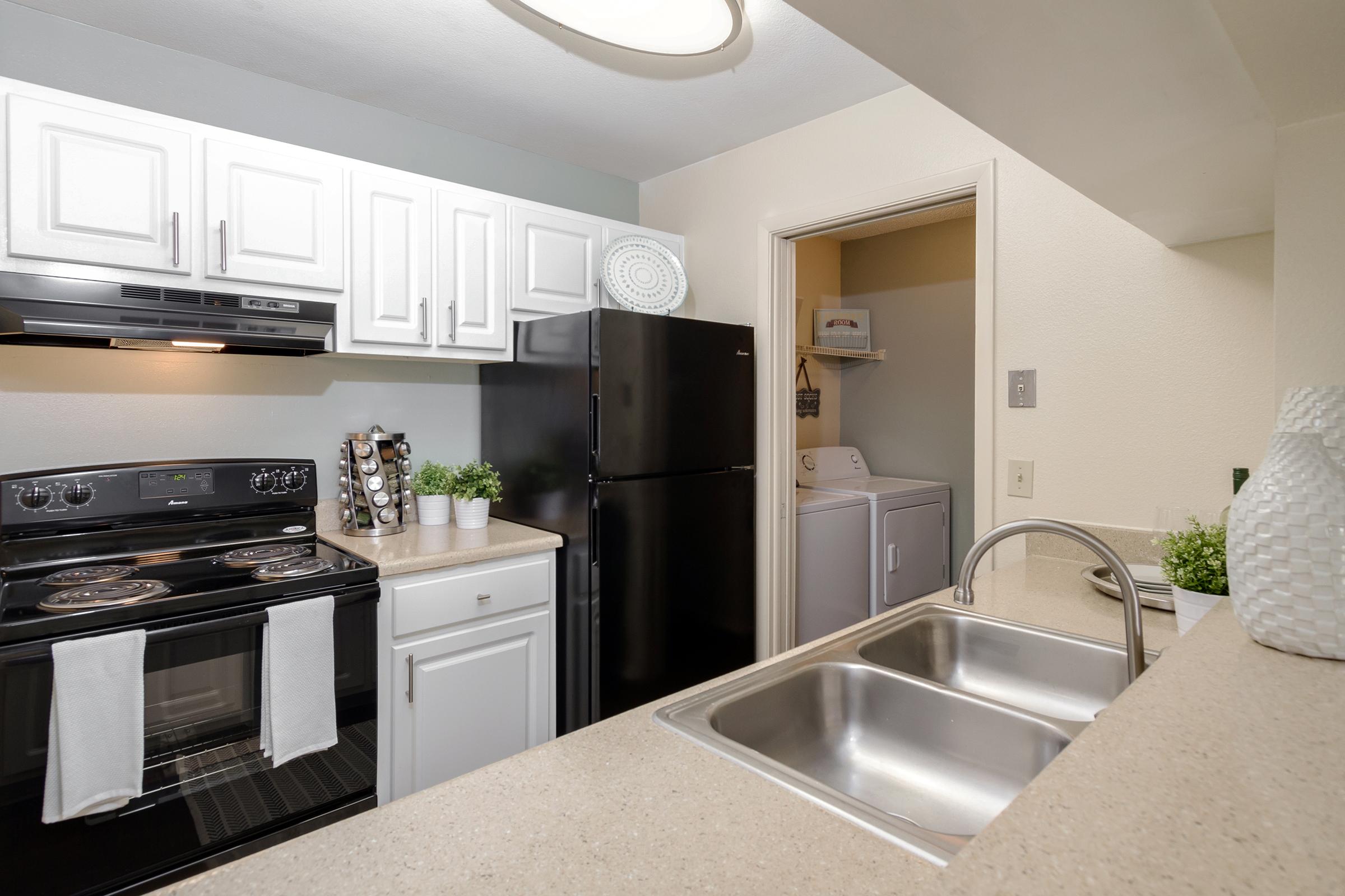 Modern kitchen featuring white cabinetry and dark appliances. Includes a black stove with a microwave above, a refrigerator, and a double sink with a stone countertop. A laundry area is visible through a door, and decorative green plants are placed on the counter for an inviting touch.