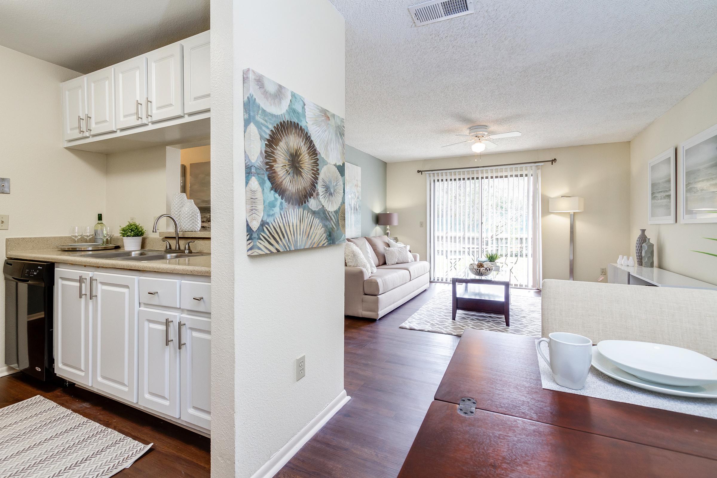 View of a modern living space featuring a kitchen with white cabinetry and a dining area with a wooden table set. The living room showcases a light-colored sofa, a ceiling fan, and large windows with natural light. Decorative art on the wall adds a touch of elegance, while a neutral color palette creates a cozy atmosphere.