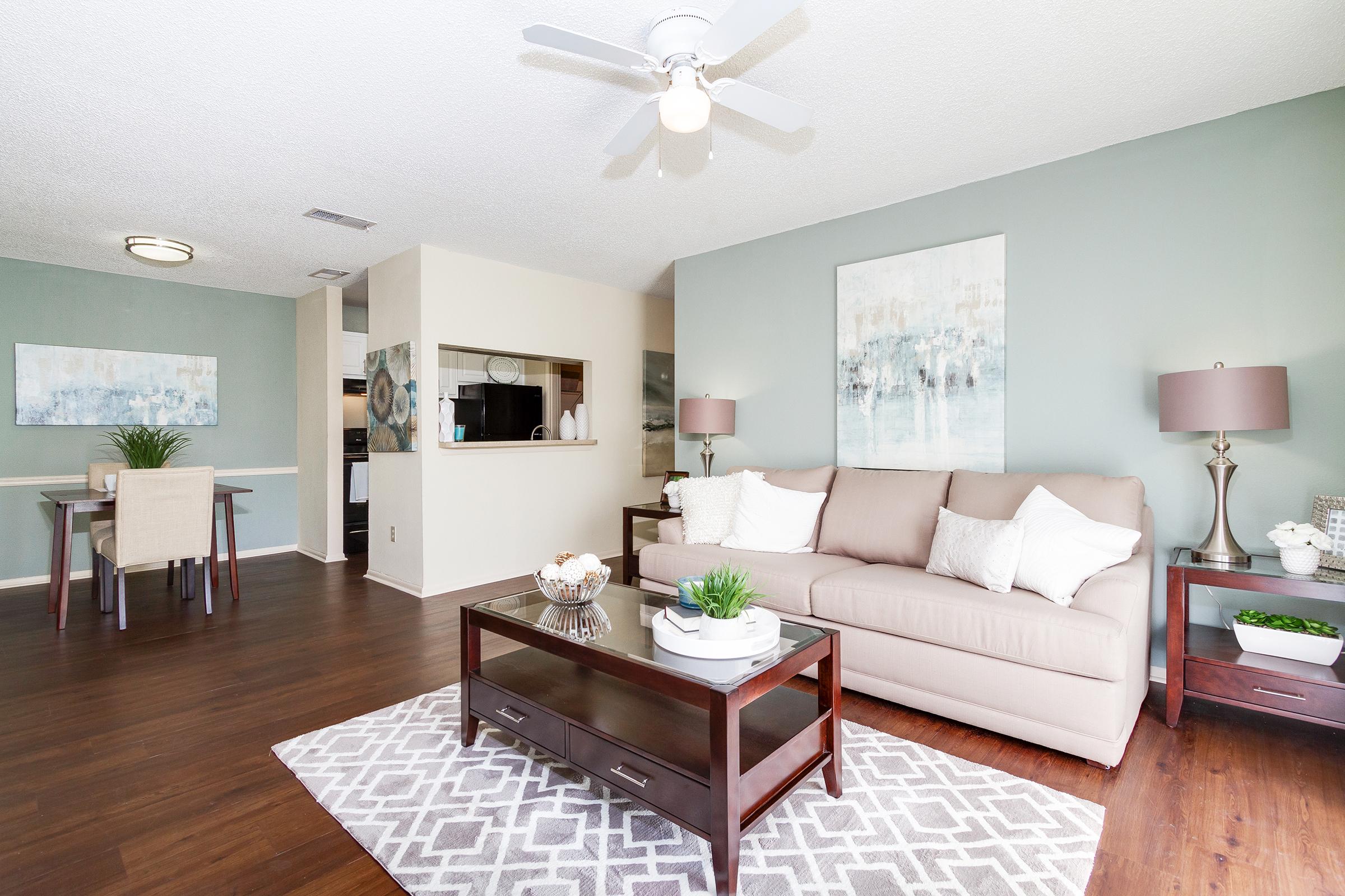 A modern living room featuring a beige sofa with white pillows, a wooden coffee table with decorative plants, and a light-colored area rug with a geometric pattern. There's a ceiling fan, and the room has a stylish paint contrast with an adjacent dining area visible in the background.