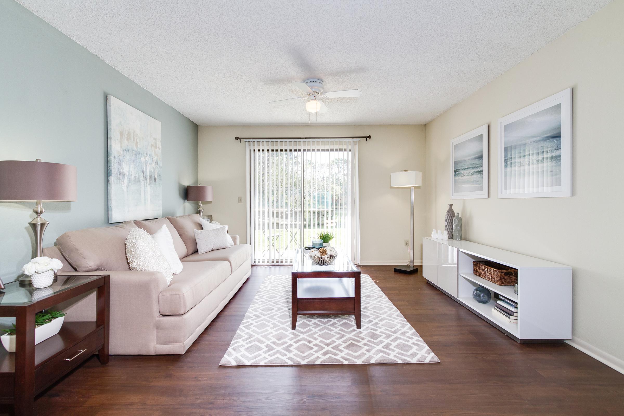 Bright and inviting living room with a light-colored sofa adorned with decorative pillows, a wooden coffee table, and a stylish area rug. The space features neutral walls, framed artwork, and a ceiling fan, with large glass doors leading to a patio area, allowing natural light to fill the room.