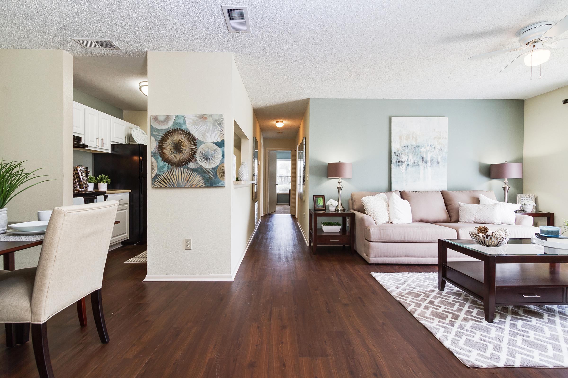 A brightly lit living room featuring a beige sofa, coffee table, and decorative artwork on the walls. A dining area with a small table and chairs is visible on the left. The space has a modern design with hardwood floors and a light color palette, creating a warm and inviting atmosphere.