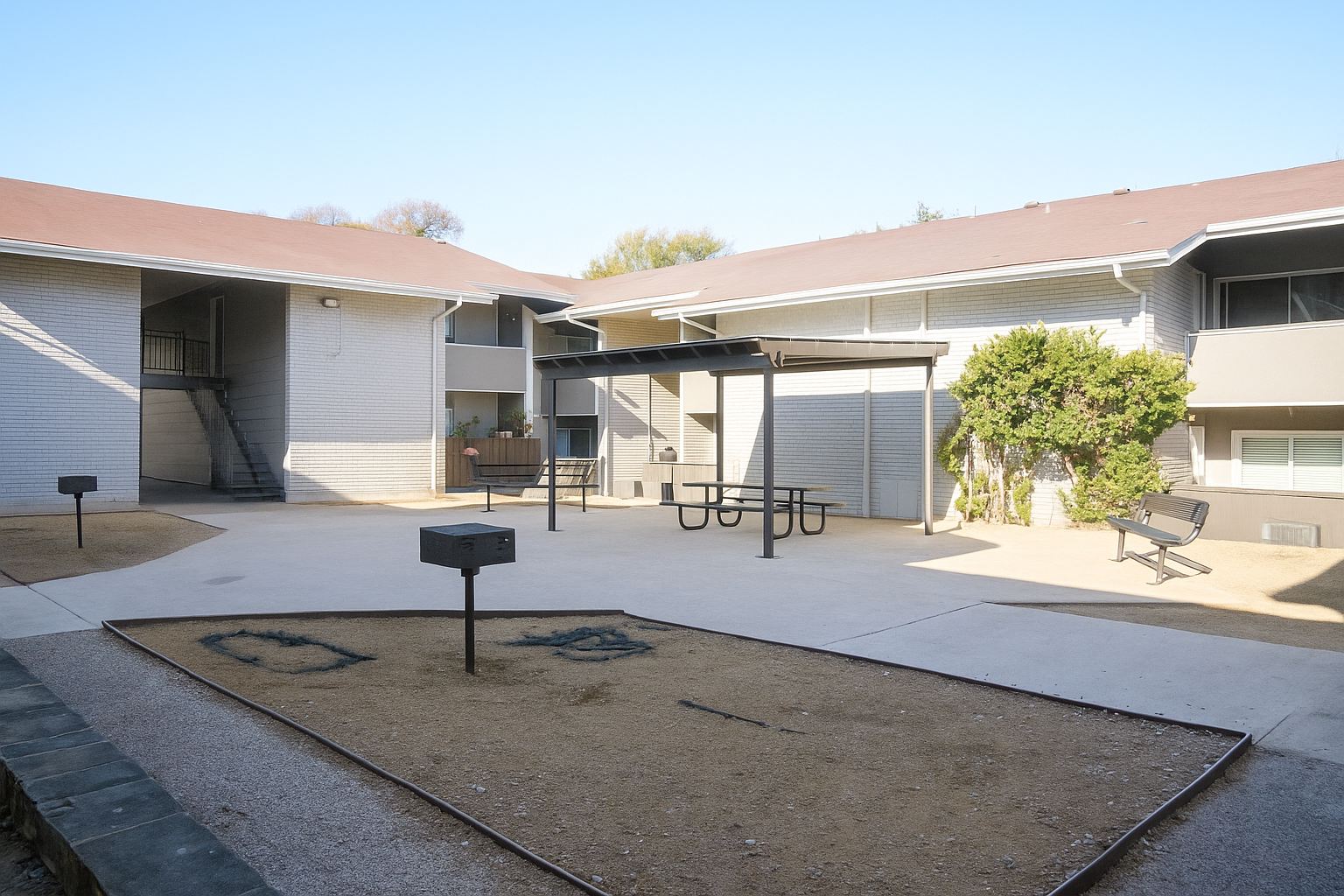 A view of a courtyard in an apartment complex featuring two adjacent buildings, a picnic table with benches, a grill, and a small grassy area. The ground is mostly paved with some sandy patches, and there are stairs leading to upper levels in the background under a clear blue sky.