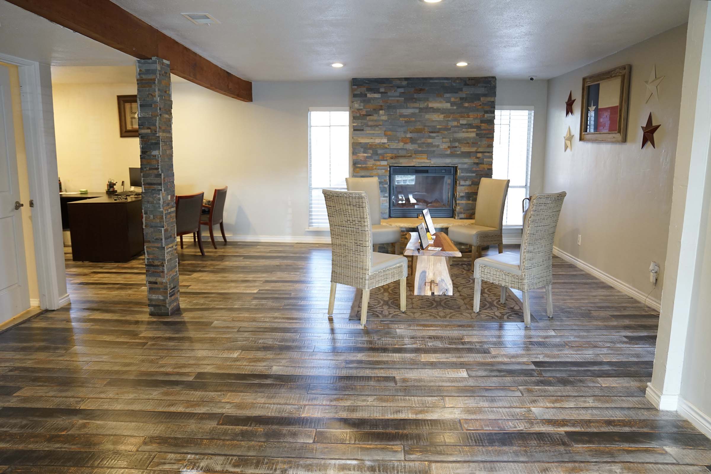 A modern interior space featuring a cozy seating area with two wicker chairs and a wooden coffee table. The walls are decorated with stone accents and stars, and a fireplace adds warmth. Light wooden floors enhance the inviting atmosphere, with a glimpse of a dining area in the background.