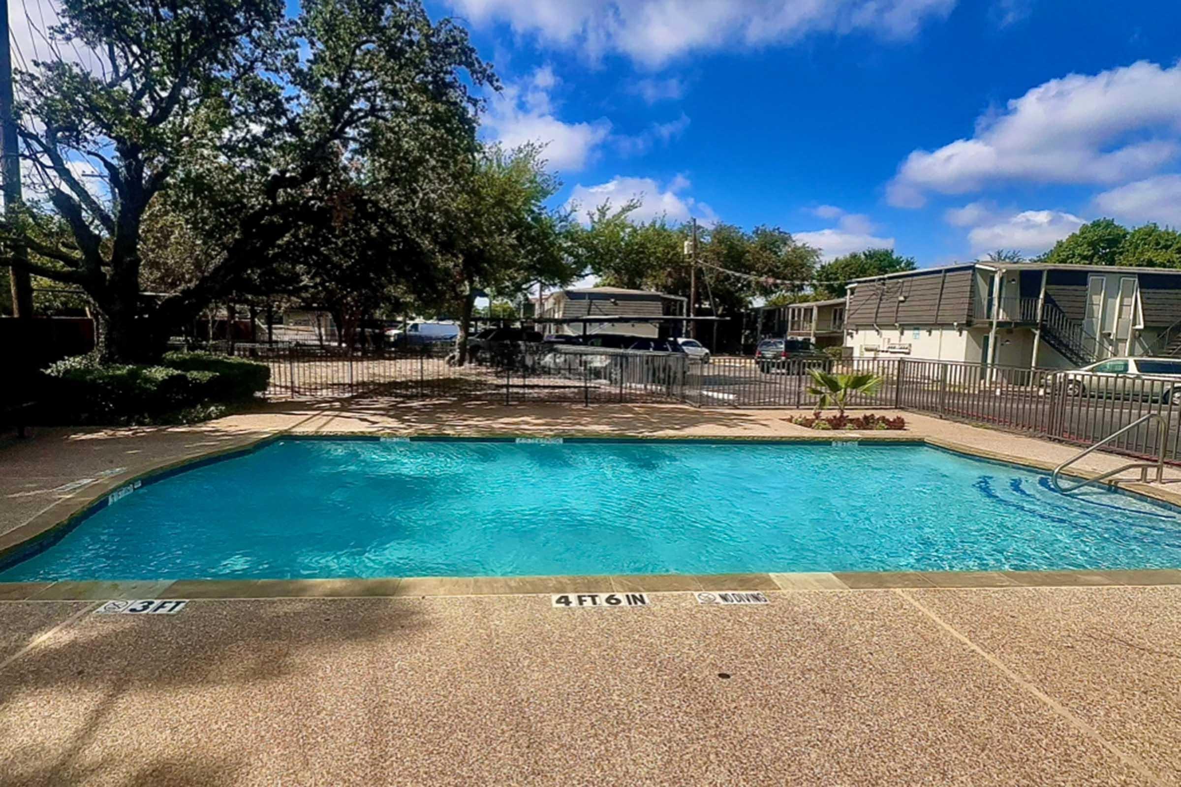 A clear blue swimming pool surrounded by a concrete deck, featuring poolside landscaping and chairs. The background includes trees, a fence, and nearby buildings under a partly cloudy sky. The depth markings of the pool are visible along the edge.