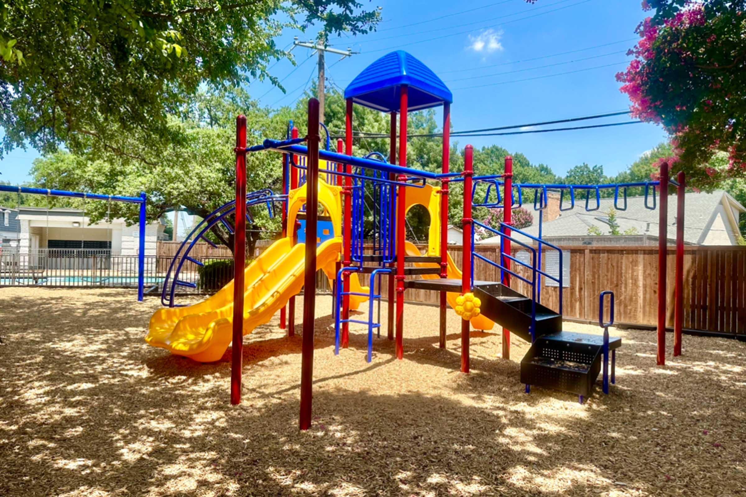 A colorful playground featuring a yellow slide, climbing structures, and various play equipment, surrounded by wood chip flooring and trees. The bright blue and red structures create an inviting space for children to play in a sunny outdoor setting.
