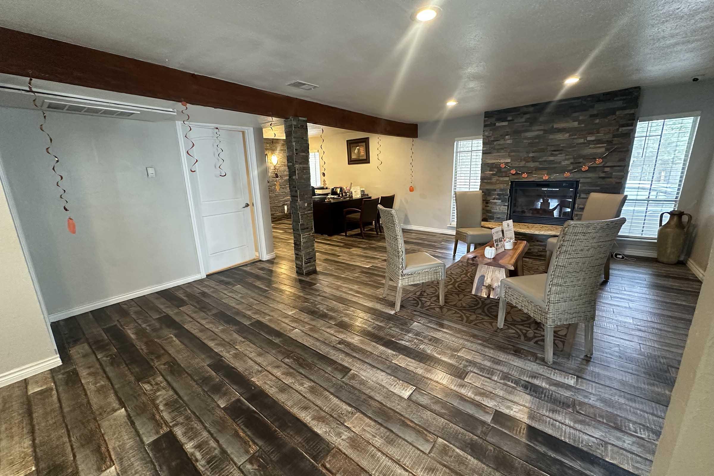 A modern interior view of a spacious living area featuring a cozy dining space with four beige chairs around a wooden table. A stone fireplace is visible on one side, and the room is decorated with orange string lights and simple decor. The flooring is made of wooden planks, providing a warm ambiance.