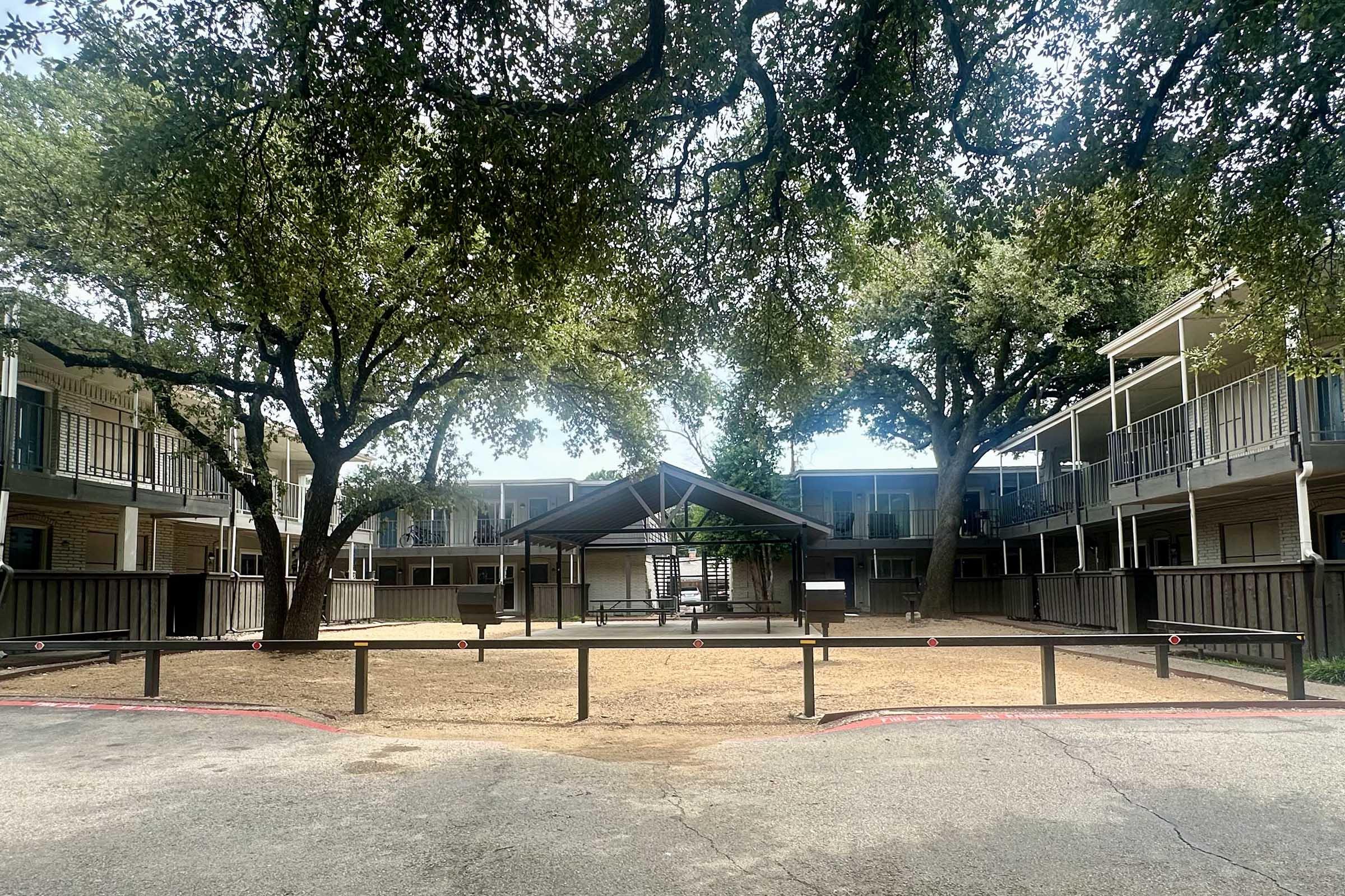 A courtyard surrounded by two-story apartment buildings, featuring a sandy area with wooden fencing and an open pavilion in the center. Large trees provide shade, and there are several balconies visible on the buildings. The scene has a serene and communal atmosphere.