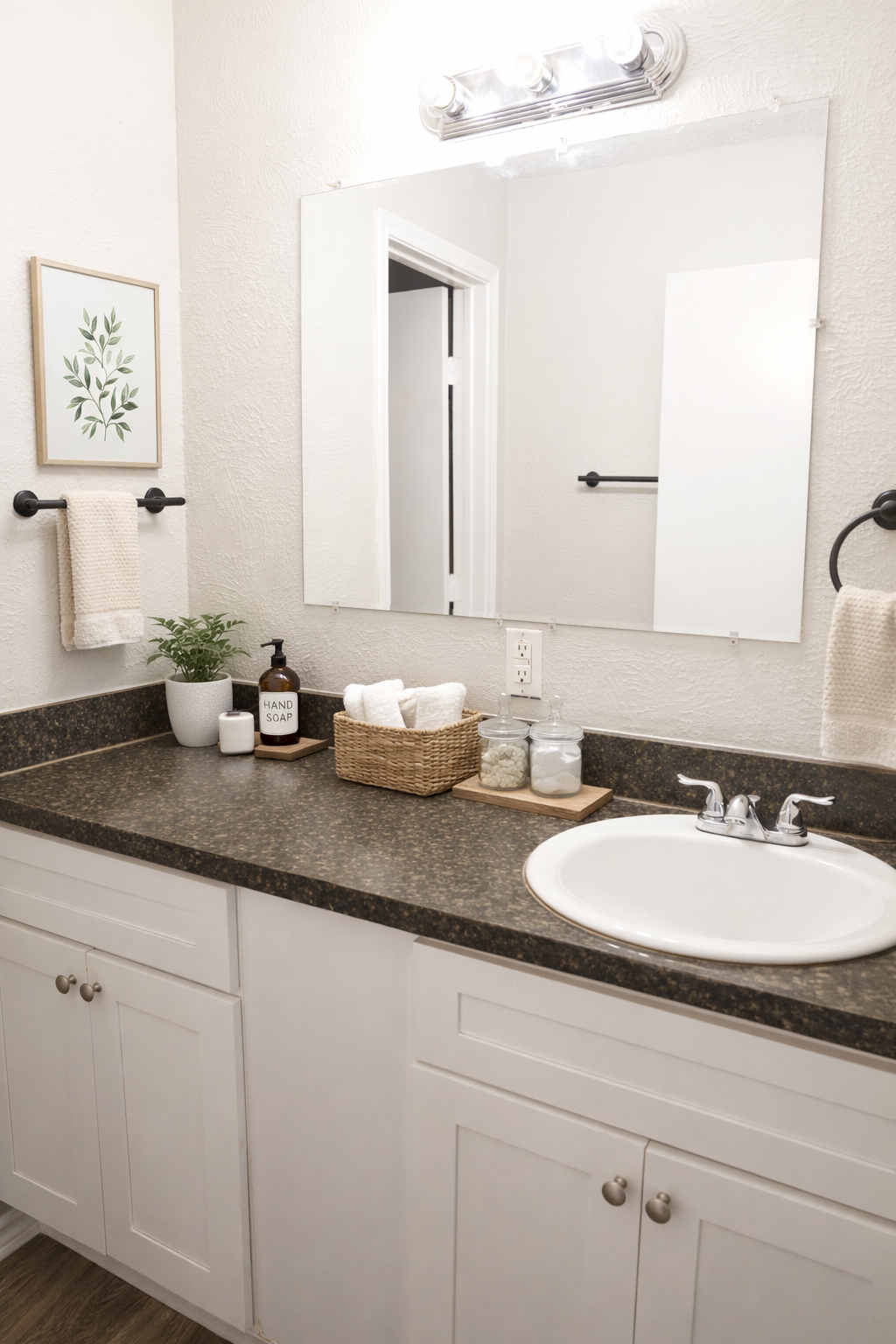 A clean bathroom interior featuring a countertop with a sink, a basket holding towels, a plant, and jars. Above the sink, there is a mirror and a light fixture. The walls are painted in neutral tones, and there are decorative elements like a framed plant print and hand soap bottle.