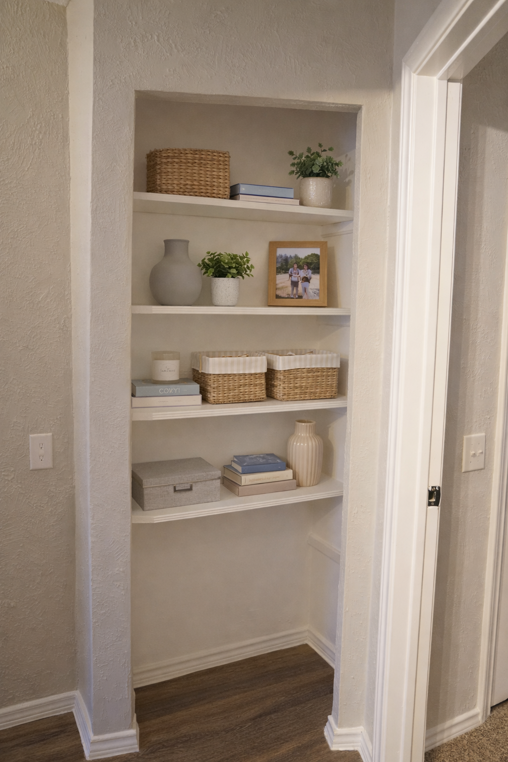 A neatly arranged small shelf nook featuring several white shelves. The top shelf displays a small plant and books, the middle shelf includes decorative boxes, and the bottom shelf holds a light-colored vase. There's a framed photo on the left and light-colored walls and wood flooring enhance the cozy atmosphere.