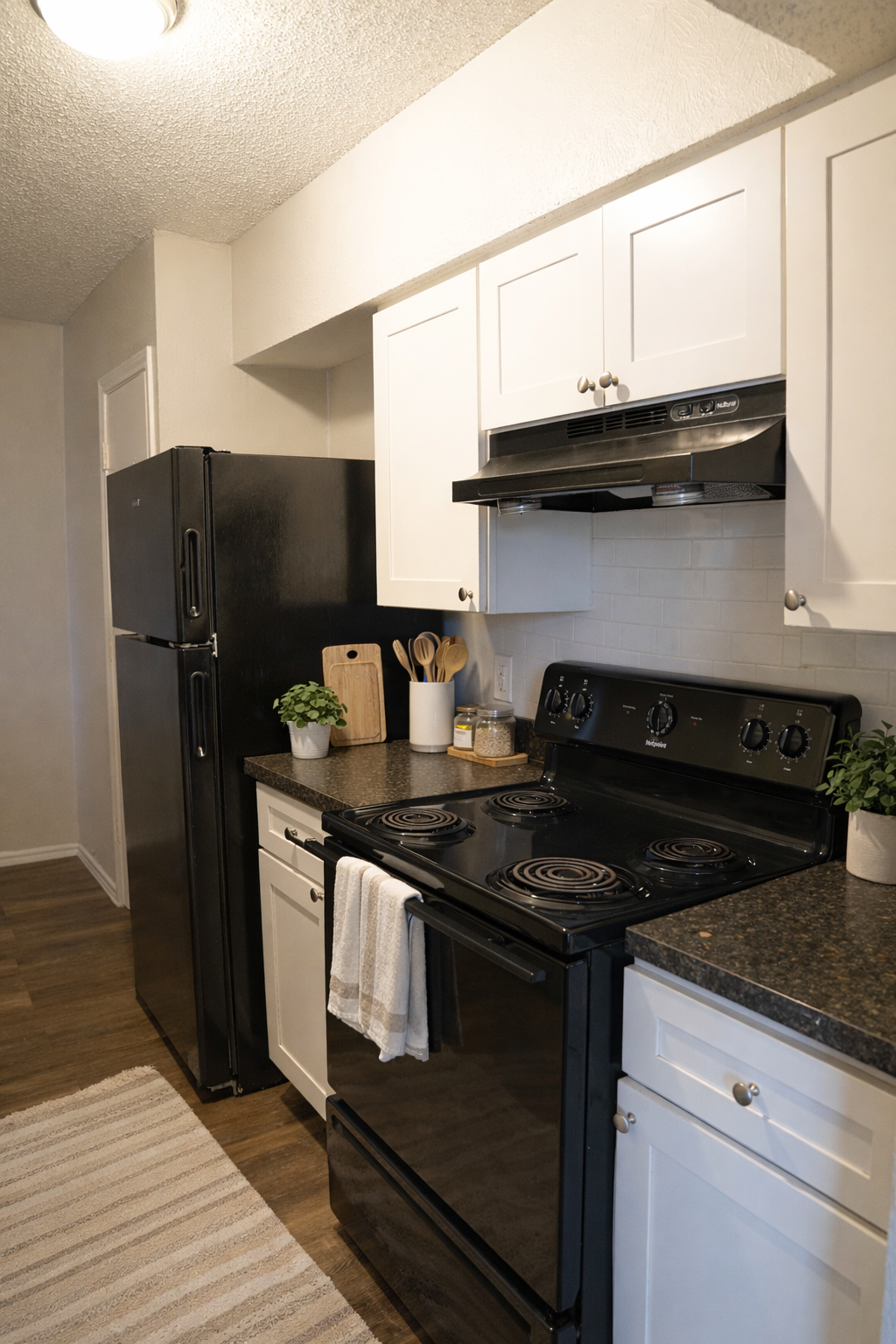 A modern kitchen featuring a black stove and refrigerator, white cabinets, and a granite countertop. The space includes cooking utensils, a wooden cutting board, and small plants for decoration. A light-colored rug is on the floor, and the walls are painted in a neutral tone.