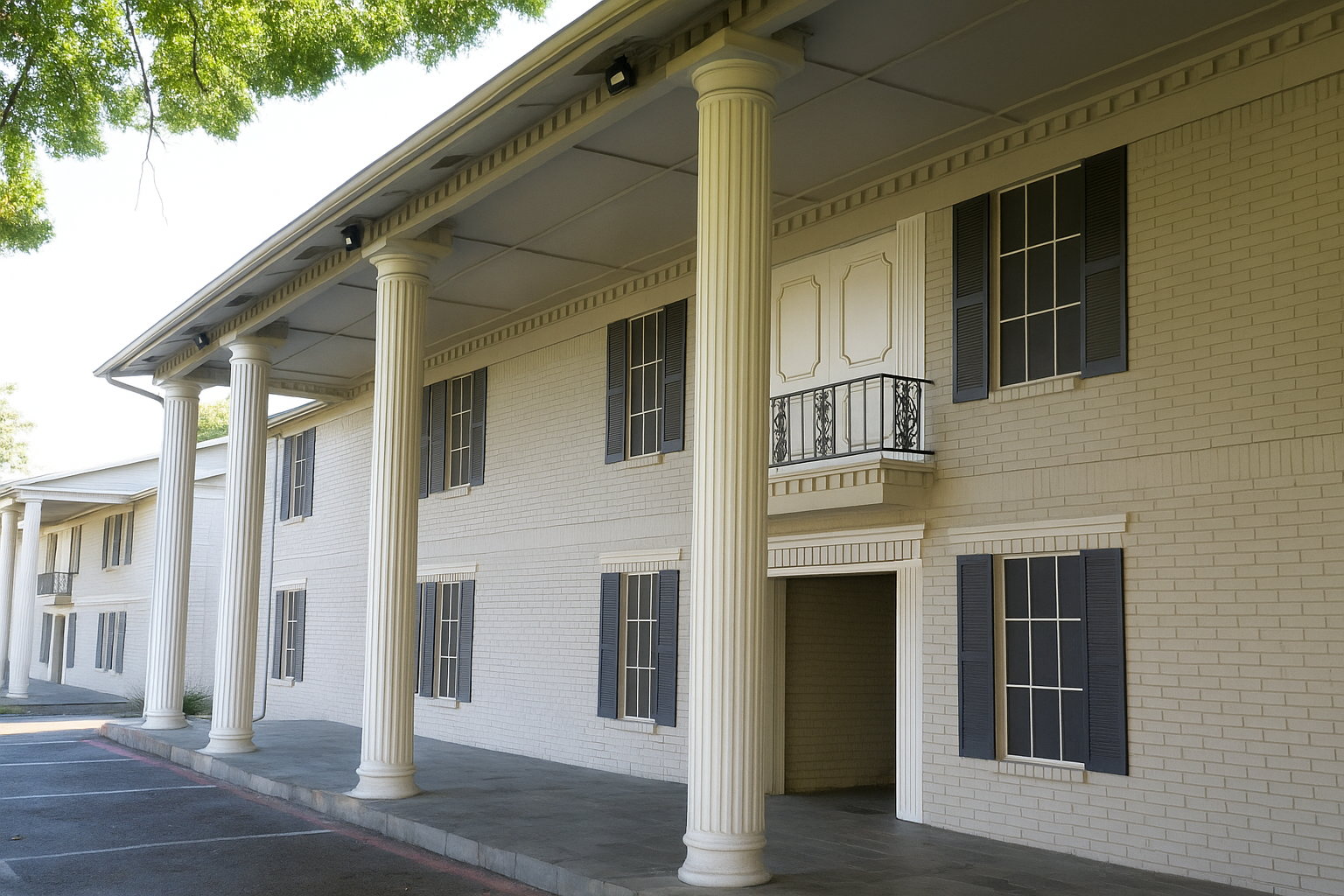 Elegant white building featuring tall columns, a covered porch area, and large windows with dark shutters. A decorative balcony with wrought iron railing is visible above the entrance. The façade is clean and modern, with a landscaped area nearby.