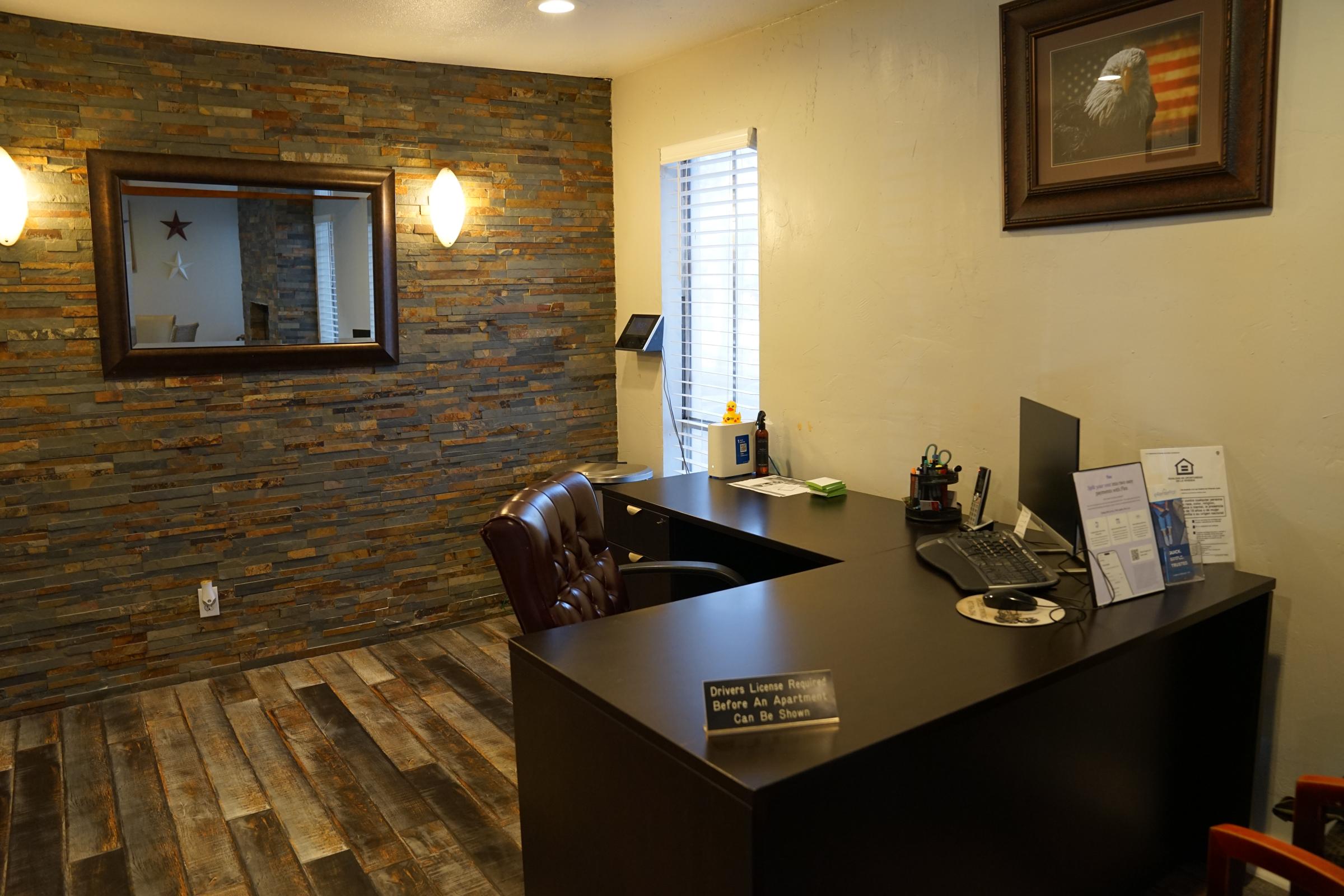 Reception area with a dark wooden desk, comfortable chair, and computer setup. The wall features a textured stone design and a large mirror, while a flag and eagle artwork is displayed. Soft lighting creates a welcoming atmosphere. A small brochure stand is on the desk.