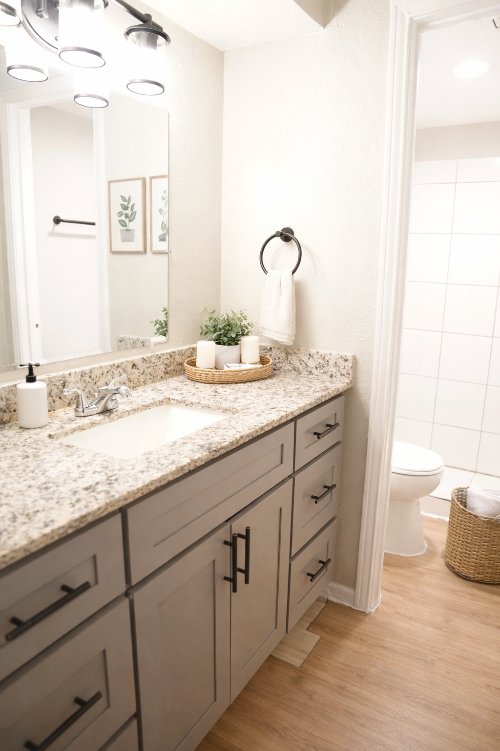 A well-designed bathroom featuring a granite countertop with a sink, modern faucet, and decorative items on a wooden tray. The walls are light, with framed botanical art. A mirror reflects the space, and a towel hangs from a rack. A white shower and toilet are visible in the background, with wooden flooring.