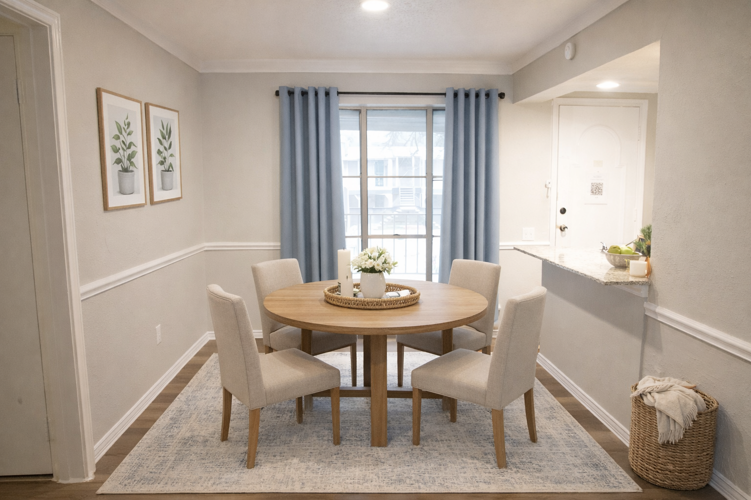 A modern dining area featuring a round wooden table surrounded by four light-colored upholstered chairs. The room has neutral walls and a light blue rug, with framed botanical prints on the wall. Large windows dressed with blue curtains allow natural light to fill the space, and a wicker basket adds a decorative touch.
