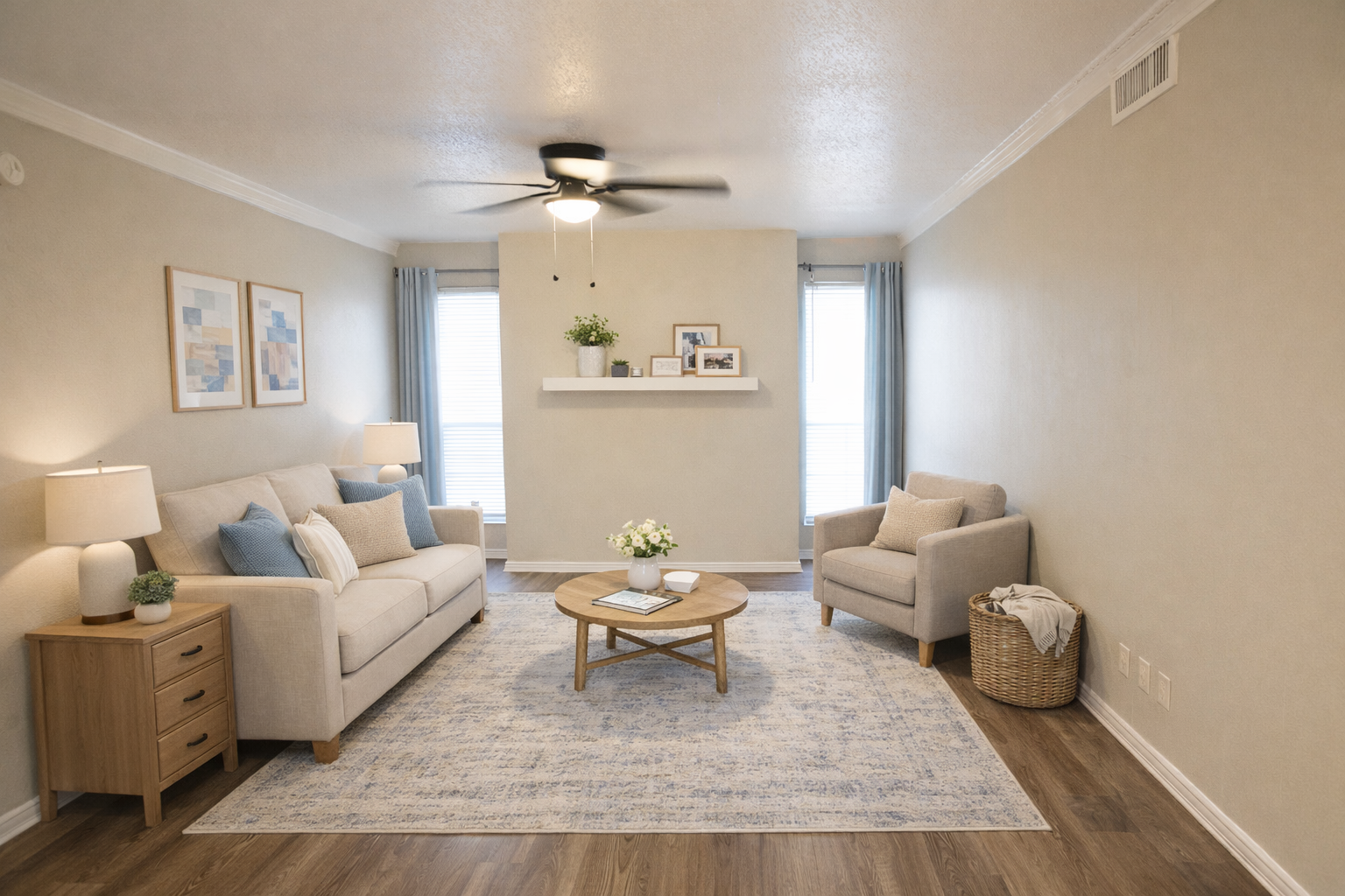A cozy living room featuring a light-colored sofa with blue and beige cushions, a round wooden coffee table, and an armchair. A decorative shelf with plants and framed pictures is mounted on the wall, complemented by two windows with light blue curtains. A soft area rug covers the wooden floor.