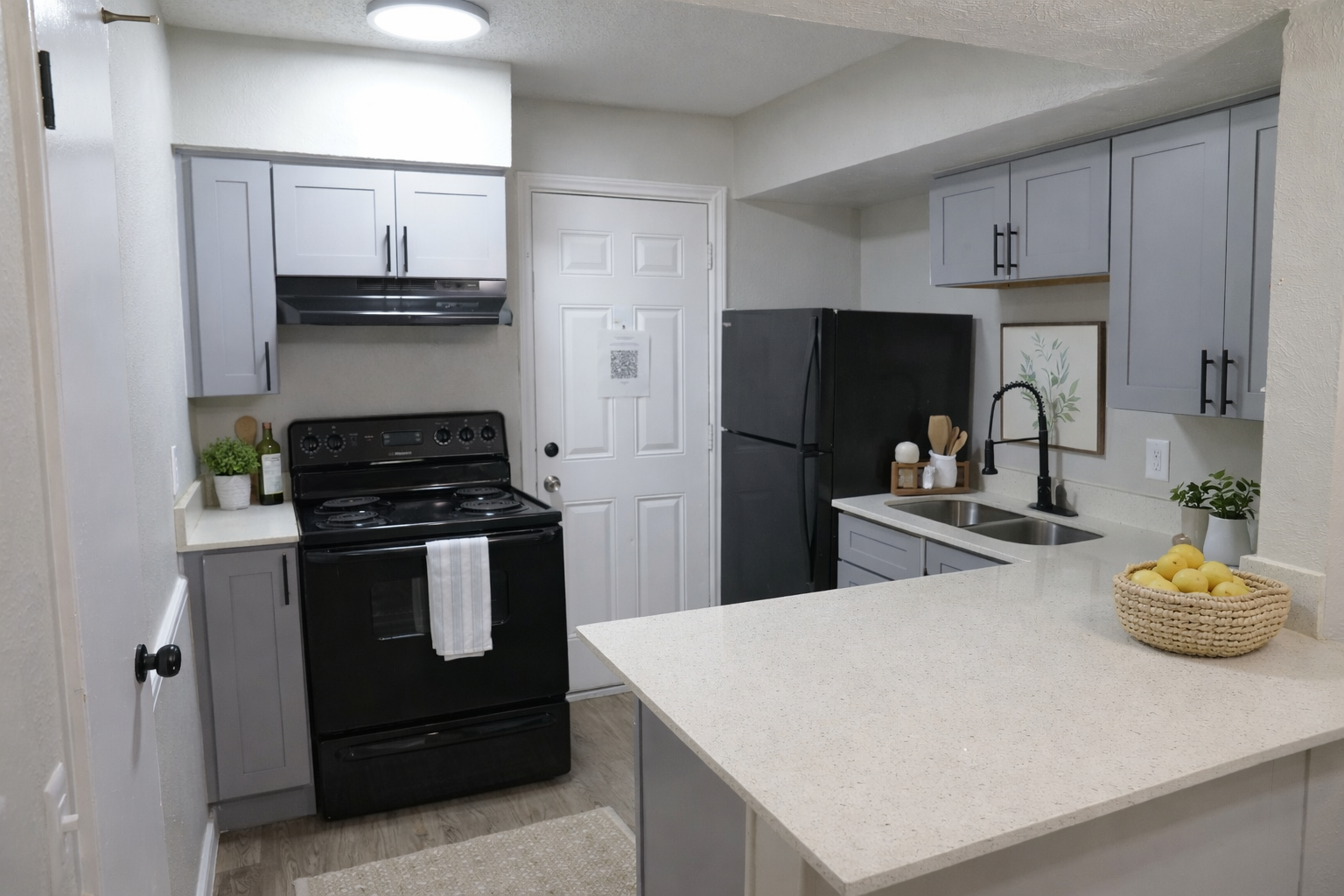 A modern kitchen featuring light gray cabinetry, a black stove and refrigerator, a sink with a faucet, and a countertop with a bowl of lemons. The space is well-lit with natural light and has a neutral color scheme, creating a clean and inviting atmosphere.