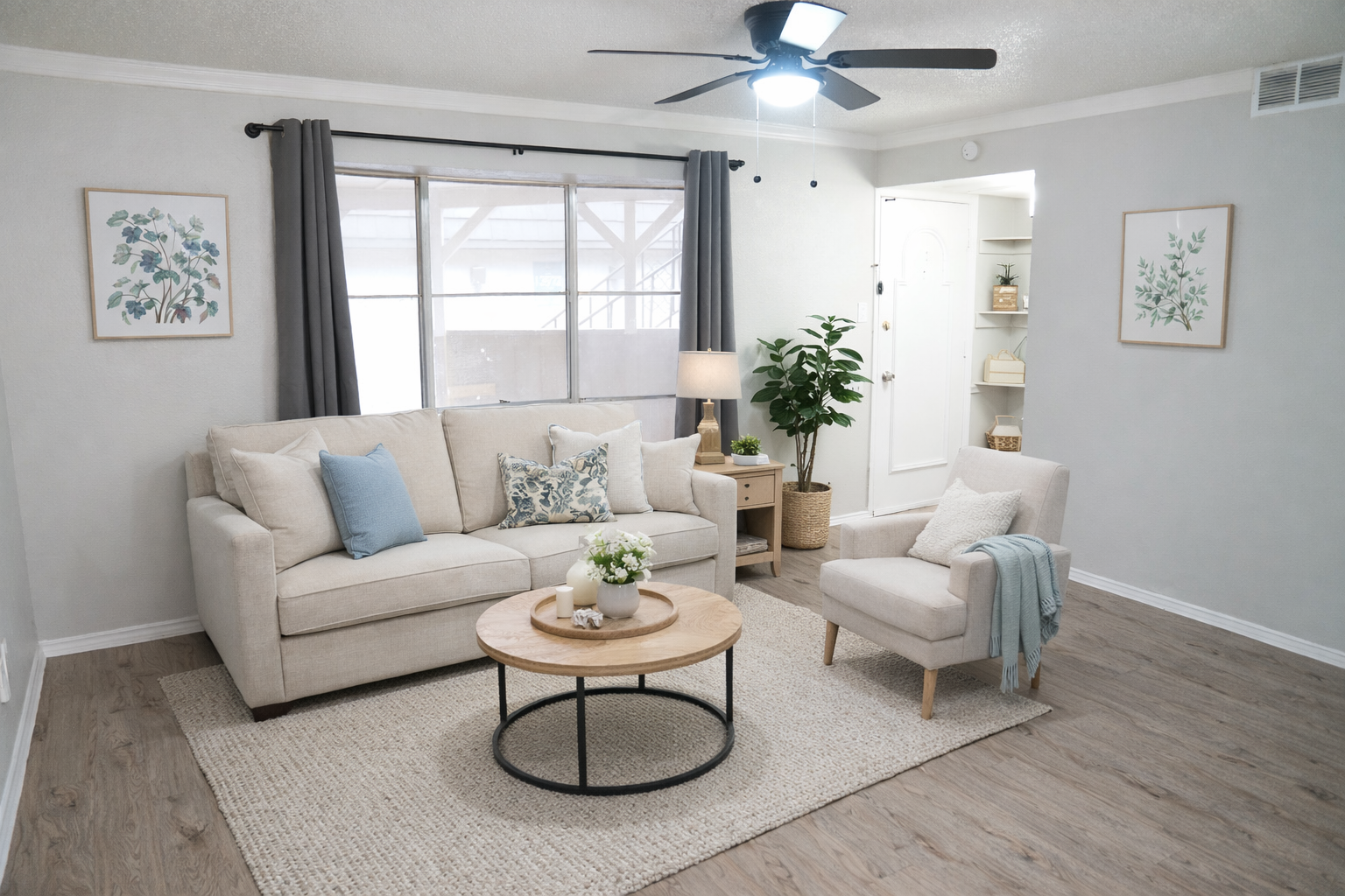 A cozy living room featuring a light beige sofa with blue and white decorative pillows, a light armchair, and a round coffee table. The space has a neutral color palette, natural wood flooring, and houseplants, with soft lighting from a ceiling fan and natural light coming through a large window.